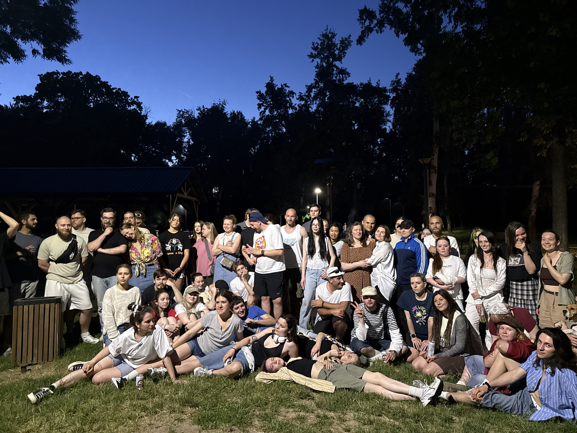 A large group of approximately 45 people is gathered outdoors in the Cukarica Urban Municipality, Serbia, possibly in a park or grassy area. The event seems to be taking place in the evening or late afternoon, as the sky is a deep twilight blue, and the lighting appears to be a mix of natural and artificial illumination.
The people are of varying ages and are dressed in casual attire. Many are standing, while others are sitting or lying on the grass. There's a relaxed and informal atmosphere, suggesting a social gathering or casual event. Some individuals are interacting with each other, and it appears that a few are posing for the photo.
In the background, there are trees and possibly a structure or covered area, which may be a pavilion or shelter. A trash can is visible on the left side of the frame. There is also a light pole. The overall impression is one of a communal gathering in a park-like setting.