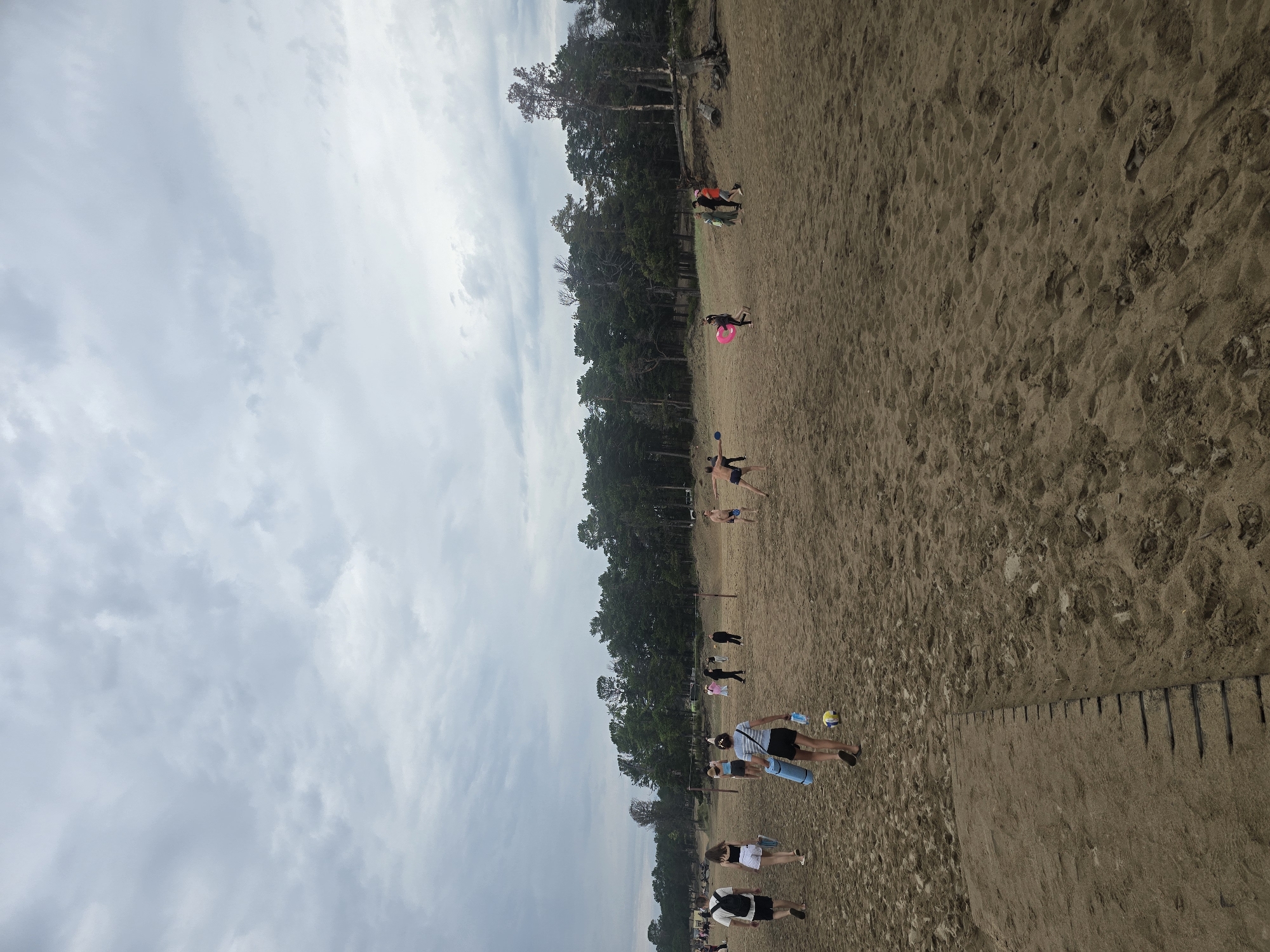 The image depicts a beach scene in Goryachinsk, Russia, with a cloudy sky overhead. The foreground consists of sandy ground with footprints. Several individuals are scattered across the beach. On the left, two people are walking towards the foreground, with one carrying a backpack. Further into the scene, a group of people are engaged in activities. Some appear to be playing volleyball, indicated by a net and a ball. Others are walking or standing. A person in the middle ground is holding a rolled-up mat and a water bottle, with a yellow ball near their feet. In the mid-right, a person is throwing a disc. In the background, a dense line of trees forms a treeline along the edge of the beach.