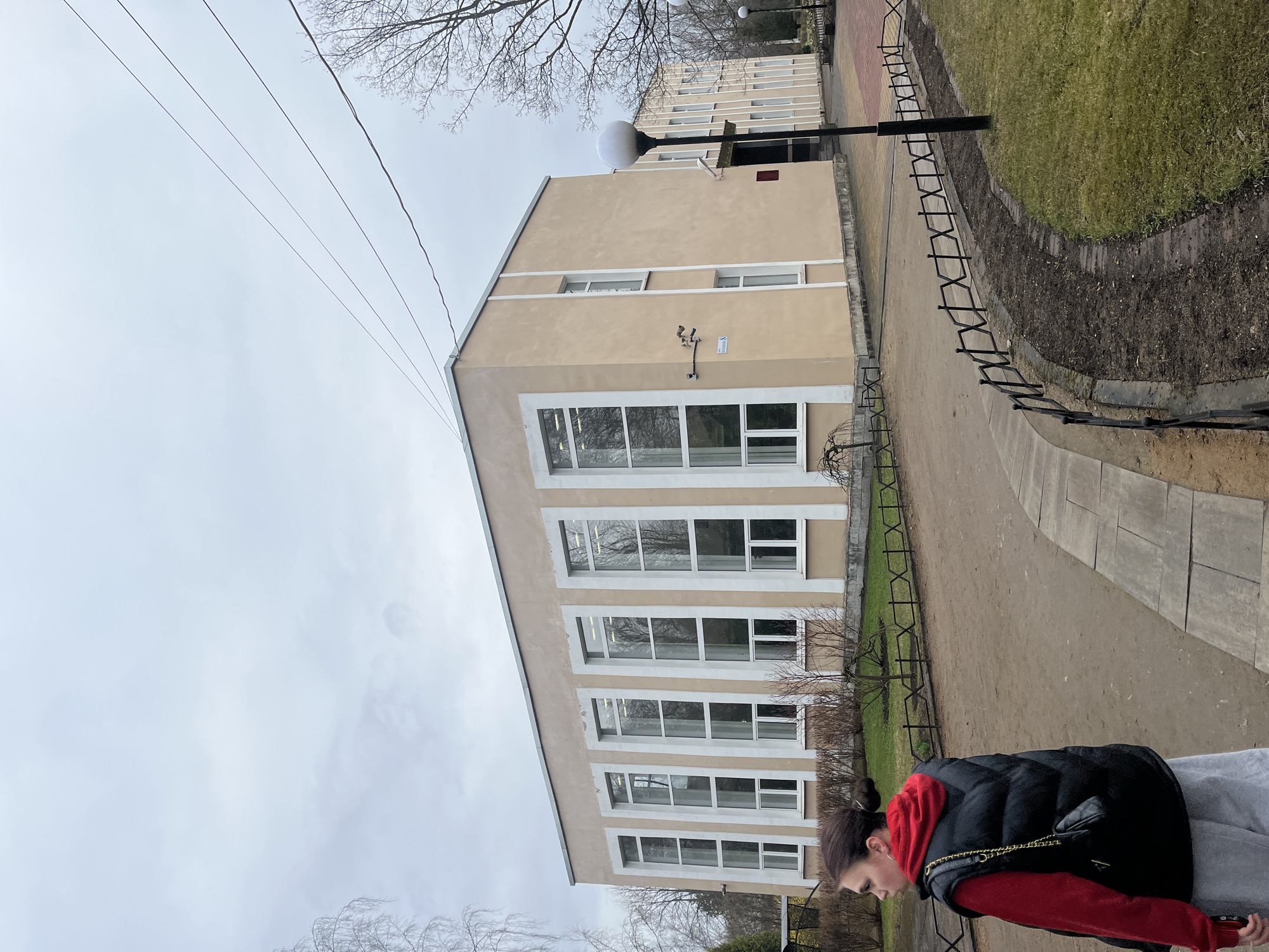The image shows a building in Saint Petersburg, Russia, likely a municipal building, taken from a slightly low angle on an overcast day. The building is light brown with white window frames. It has multiple floors with large windows. A black decorative fence runs along a walkway next to the building. A person with dark hair pulled back and wearing a red hooded jacket with a black vest and light grey pants is partially visible in the foreground. Power lines stretch across the sky. A grassy area with trees is present behind the fence.