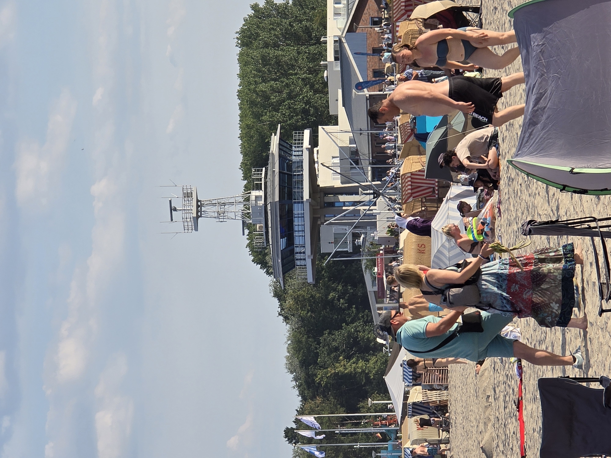 The image captures a lively scene on a sandy beach, likely in Lübeck, Germany, given the presence of a prominent maritime control tower. In the foreground, several beach chairs and tents are set up, indicating a day of leisure. A couple is walking on the sand, the man in a turquoise polo shirt and shorts, and the woman in a flowing, colorful skirt and carrying a backpack. Other individuals are scattered across the beach, some sitting in chairs, others walking or playing.
In the background, the striking maritime control tower stands tall, its modern design contrasting with the natural beach environment. The tower features multiple levels with large glass windows and is topped with radar equipment. Behind the tower, a line of trees forms a green backdrop, and further back, buildings can be seen. Flags are visible flying on poles near the beach structures. The sky is partly cloudy, with a few white clouds against a bright blue expanse. The overall atmosphere appears to be one of a pleasant summer day at the beach.