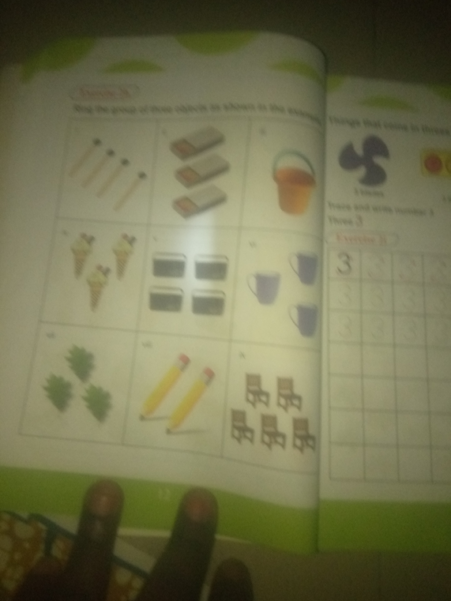 The image shows a page from a children's educational book, likely a workbook focused on counting or numbers. The page is divided into sections with different illustrations. On the left side, there are rows of objects: matches, ice cream cones, green leaf-like shapes, and what appear to be pencils. In the top right section, there is a drawing of a fan, and the text "Things that come in threes". Below that, there is an exercise labeled "Trace and write number 3". To the right of this is a grid for tracing the number 3. On the bottom right, there are several chairs depicted. The image is blurry and partially obscured by fingers in the foreground, indicating it was taken by a person holding the book. The environment appears to be indoors, possibly a home or classroom, with soft lighting. There is no information in the image to suggest a specific location in Jauro Yinu, Nigeria.