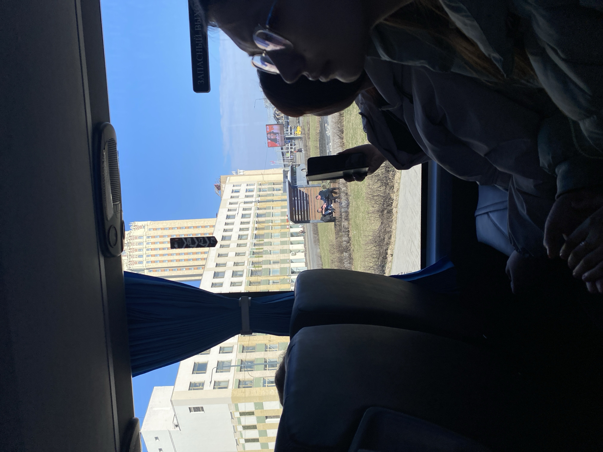 The image depicts a person inside a bus, holding a smartphone. The view outside the bus window shows a clear blue sky and several buildings in Moscow, Russia. One prominent building is a large, rectangular structure with multiple windows, cream and brown in color, identifiable as Hotel Kosmos. In the distance, a billboard is visible, and a few people are sitting on a bench in a park-like area. The interior of the bus includes seats with blue curtains along the windows. A sign is visible in Russian, likely indicating an emergency exit.