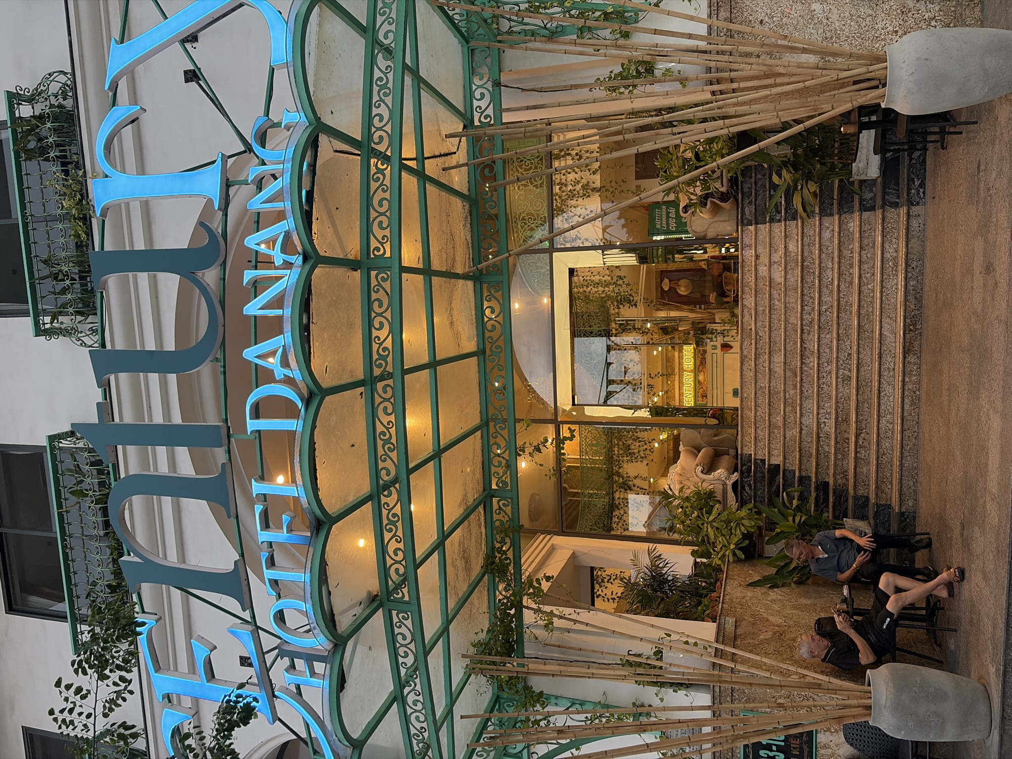 The image shows the entrance to the Century Hotel in Đà Nẵng, Vietnam. A large, illuminated sign in blue letters above the entrance reads "CENTURY HOTEL DANANG". The entrance is accessed via a wide, marble staircase leading up to a glass-enclosed lobby. In front of the entrance, several tall bamboo stalks are arranged in a large grey planter. Two men are seated on chairs to the left of the stairs, one looking at his phone and the other talking to him. The hotel facade is adorned with greenery, and the overall scene suggests an urban environment.
