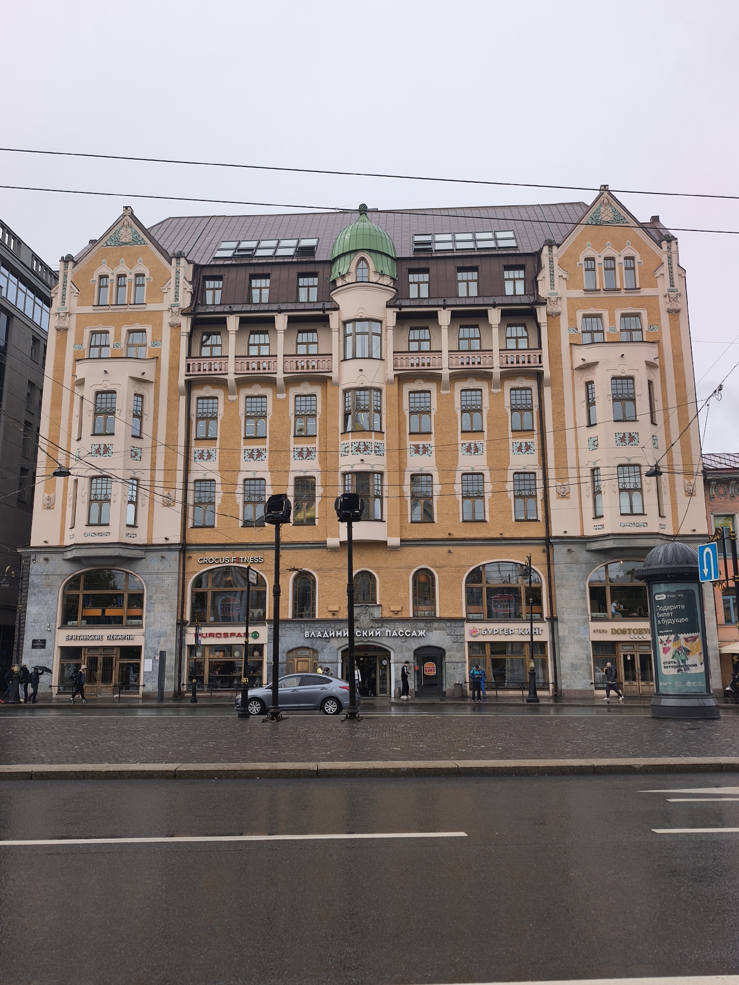 The image depicts a multi-story building with a distinctive architectural style, featuring a mix of orange and cream-colored facades, numerous windows, and a green dome atop a central tower. The building houses several commercial establishments, as evidenced by the storefront signs reading "Британские Пекарни" (British Bakeries), "EUROSPA," "ВЛАДИМИРСКИЙ ПАССАЖ" (Vladimirsky Passage), "БУРГЕР КИНГ" (Burger King), and "ОТЕЛЬ DOSTOEVSKY" (Hotel Dostoevsky).
Several pedestrians are present on the sidewalk in front of the building. A gray car is parked along the curb. A cylindrical advertising pillar stands on the right side of the frame, displaying a poster in Russian.
The road in the foreground is wet, indicating recent or ongoing rain. White lines are painted on the street. Overhead power lines are visible against a gray, overcast sky. The location is likely in a city in Russia, given the Russian signage.