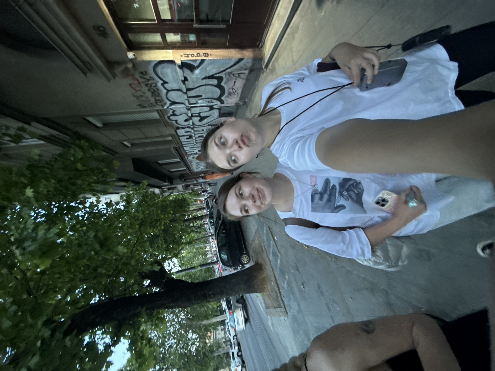 Two young women are in the frame, likely taking a selfie, in Belgrade, Serbia. They stand on a sidewalk next to a street lined with parked cars and trees. The background includes a building with graffiti. The woman closer to the camera is wearing a white t-shirt.
