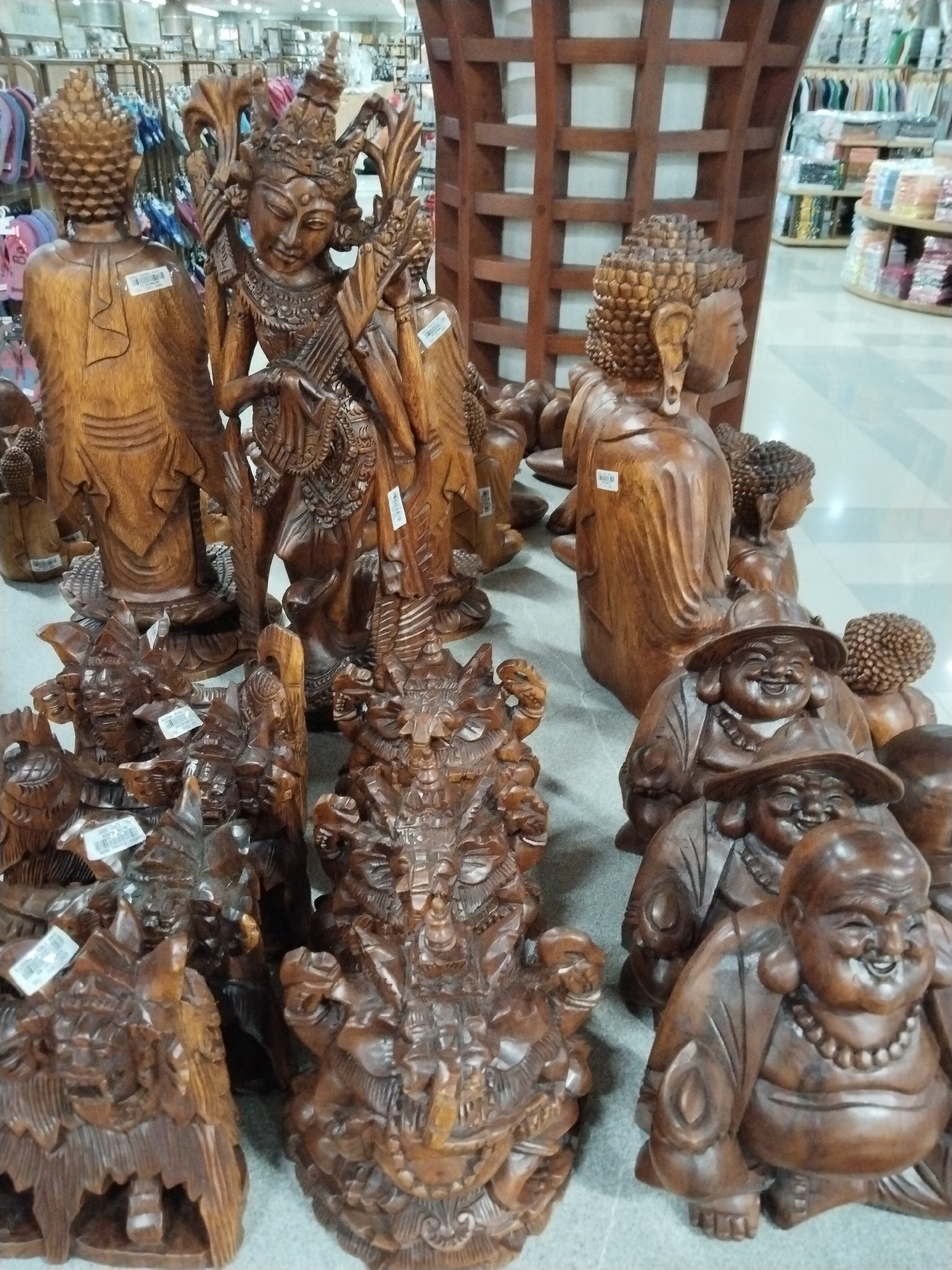 In Kuta, Indonesia, an indoor market stall displays a collection of carved wooden figurines. In the foreground, three carved "Buddha" figures, or representations of a plump, smiling man, are prominently featured. Two of these figures wear hats. Behind them, a more intricately carved figure depicts a mythical creature or deity. Further back, a large, elaborate carving of what appears to be a traditional Balinese dancer or goddess stands between two simpler wooden Buddha statues. Several other smaller wooden carvings are also visible in the background, alongside hanging clothing and shelves stocked with goods. The lighting suggests an indoor environment, possibly a shop or market.