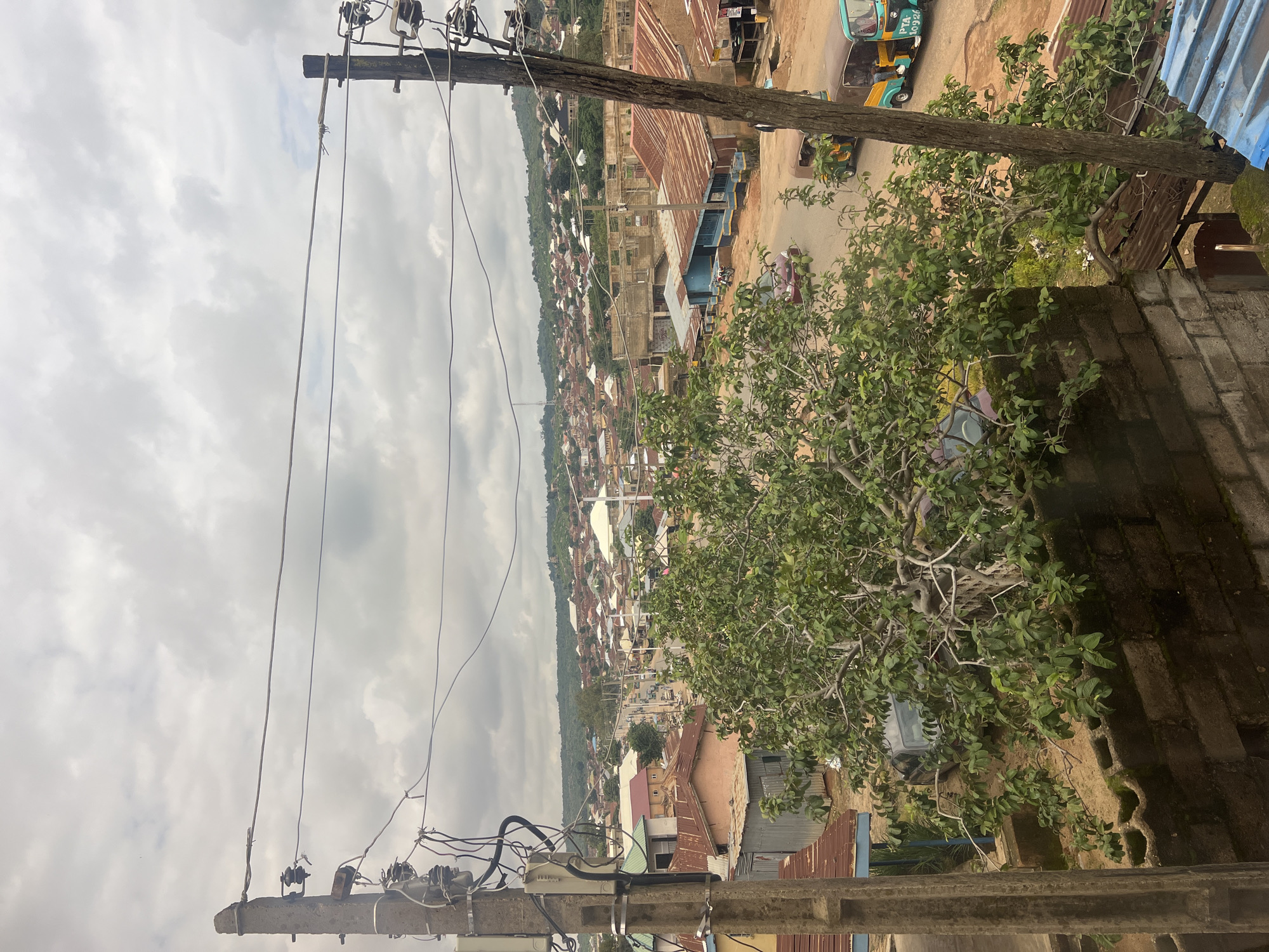 This image captures a view of a bustling street in the city of Jos, Nigeria, under a cloudy sky. The foreground is dominated by a utility pole on the left and another on the right, both adorned with electrical wires and insulators. Between these poles, a lush green tree with twisted branches spreads its foliage. In the mid-ground, a street scene unfolds with several vehicles, including a red car, two tricycles (locally known as "keke napep"), and a motorcycle. Buildings with corrugated iron roofs line the street, some appearing to be shops or homes. The background reveals a densely populated residential area with houses stretching up a gentle hillside, all under a grey, overcast sky. The overall impression is of a vibrant, active urban environment.