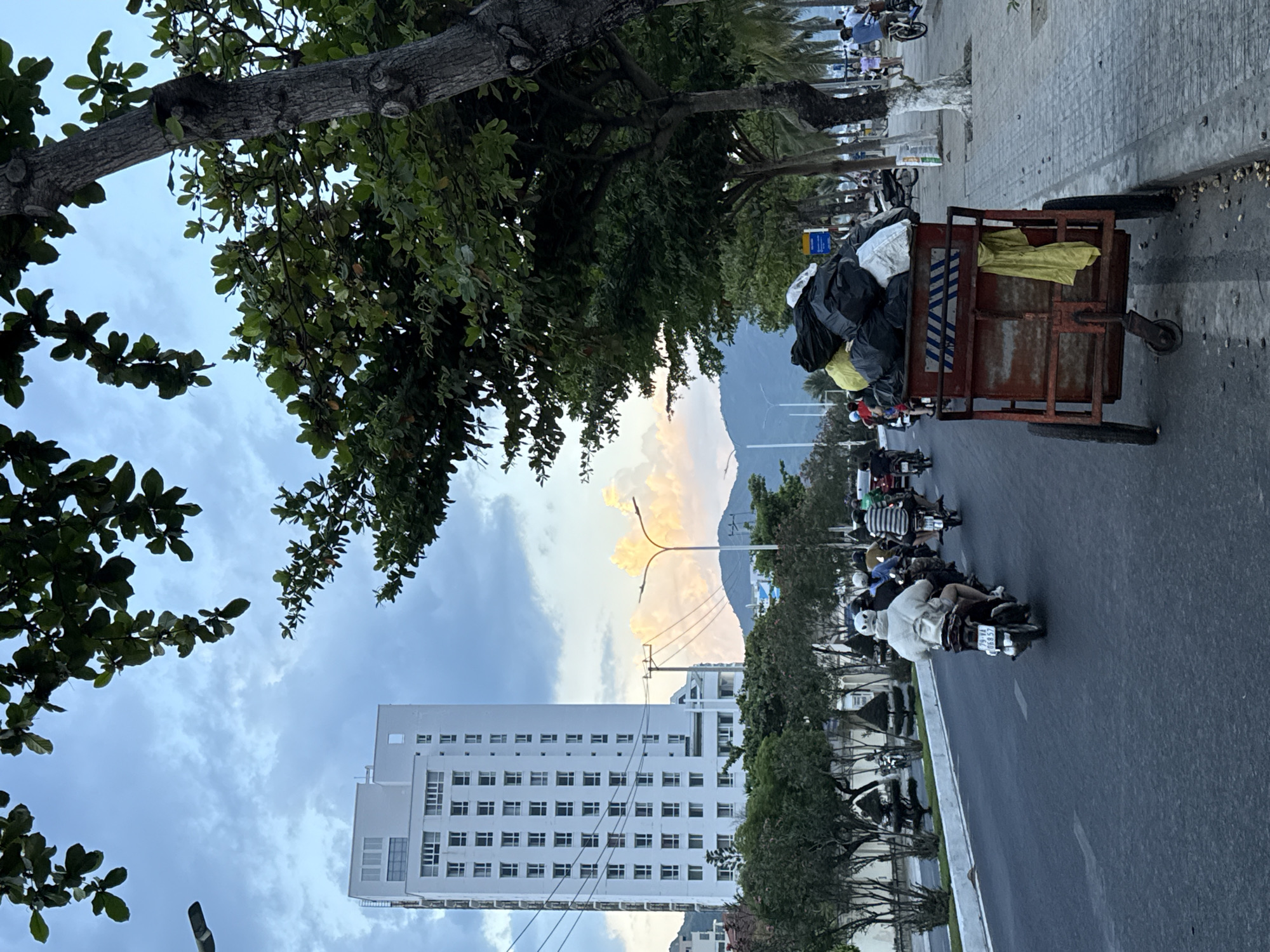 The image depicts a street scene in Bac Nha Trang Ward, Vietnam, characterized by a mix of urban infrastructure, natural elements, and daily life. A tall, white, multi-story building with numerous windows dominates the left side of the frame, standing against a cloudy sky. In the foreground, a red, two-wheeled cart filled with black and yellow trash bags is being pushed along the side of the road. Several people are riding motorbikes on the asphalt road, with some appearing to be in traffic. Lush green trees with dense foliage line the right side of the street, partially obscuring the view of the sky and the distant mountains in the background. The sidewalk is paved with gray tiles and scattered with debris. A street lamp with an orange glow is visible on the right. The overall impression is of a bustling, transitional urban environment with a blend of modern construction and natural greenery, set against a backdrop of a potentially developing landscape.