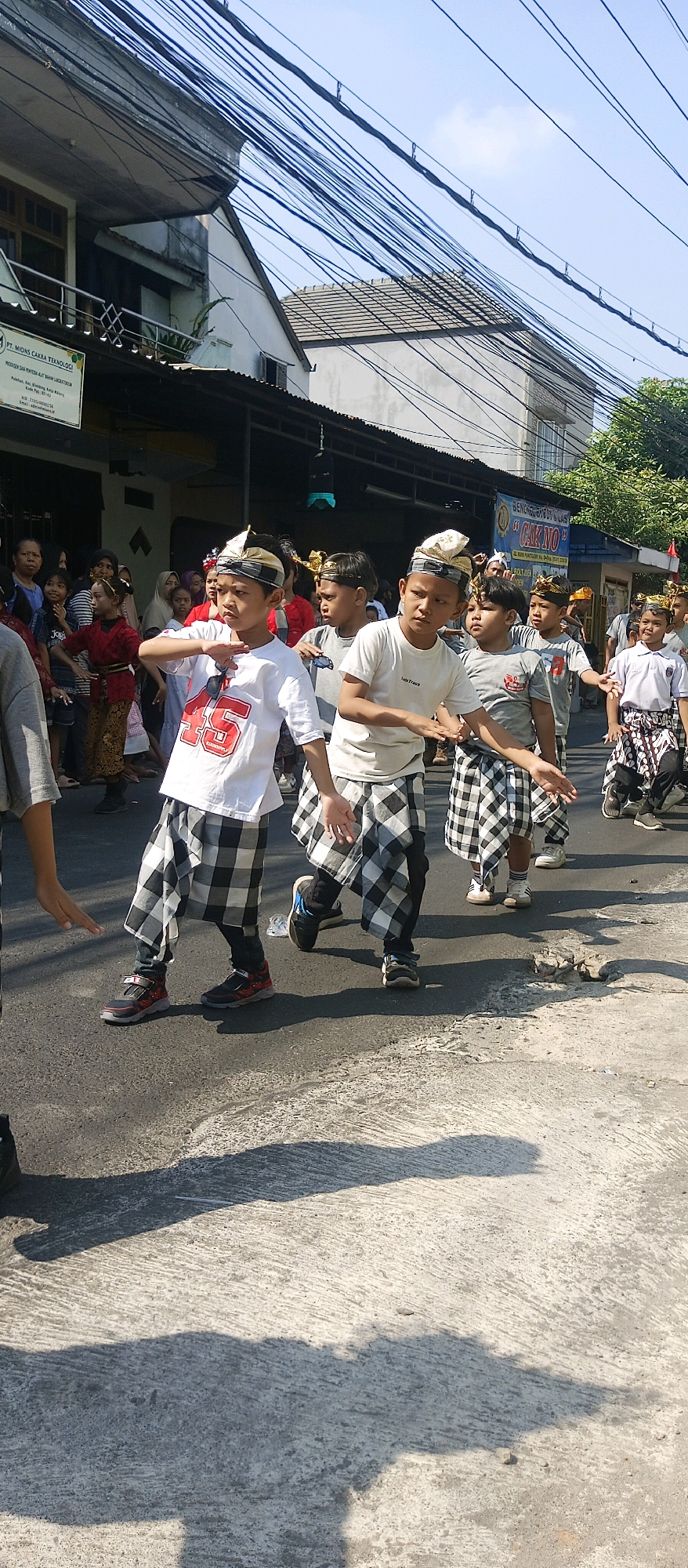 In Malang, Indonesia, a group of children are participating in a parade, likely a cultural or traditional event. The children are dressed in festive attire, featuring black and white checkered sarongs, white t-shirts with graphics, and traditional headwear. They are marching on a paved road under a clear blue sky, with power lines stretching across the frame overhead. Buildings line the street, some with corrugated metal roofs, and a sign is visible that reads "PT. MIONS CAKRA TEKNOLOGI." Spectators can be seen in the background, some watching the procession. The scene is illuminated by bright sunlight, casting distinct shadows on the road.