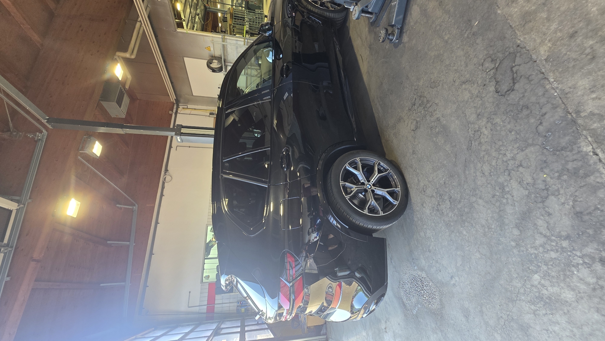 A black BMW SUV is parked in what appears to be a garage or workshop. The vehicle is angled, with its rear wheel prominently displayed. The floor is concrete and shows signs of wear and some damp patches. Above, fluorescent lights illuminate the space, and parts of the ceiling structure, including wooden beams and metal supports, are visible. To the left of the car, there are glimpses of shelving and equipment, suggesting a functional workspace. The overall lighting is bright and artificial. No people are visible in the image. The time of day and weather are not discernible. There is no visible text in the image.