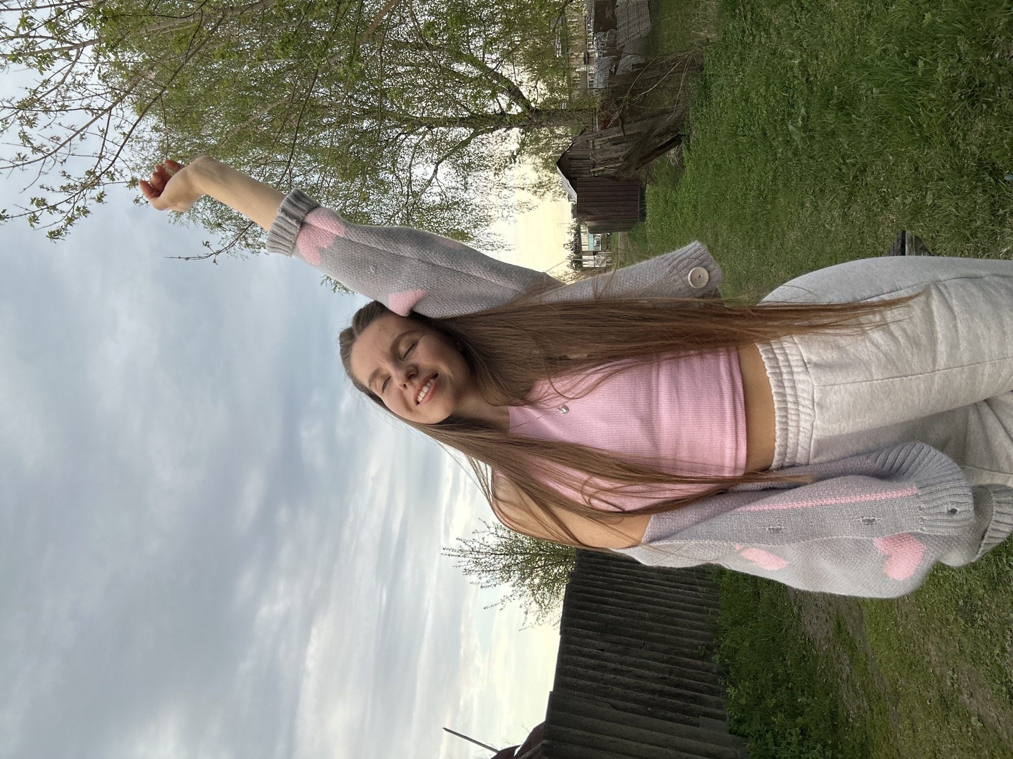 A young woman stands outdoors in what appears to be a rural or semi-rural setting, potentially within the area of Борщевка Малая, Russia, given the prompt's instruction. The woman is wearing a pink tank top, a gray sweater with pink heart designs, and light-colored sweatpants. Her right arm is raised towards a tree, while her left arm is bent at the elbow. She is smiling with her eyes closed. The environment includes a cloudy sky, a grassy area, trees, and some wooden structures, potentially sheds or outbuildings. There also appears to be a corrugated metal roof in the foreground. The image suggests a natural outdoor setting, possibly a garden or yard.