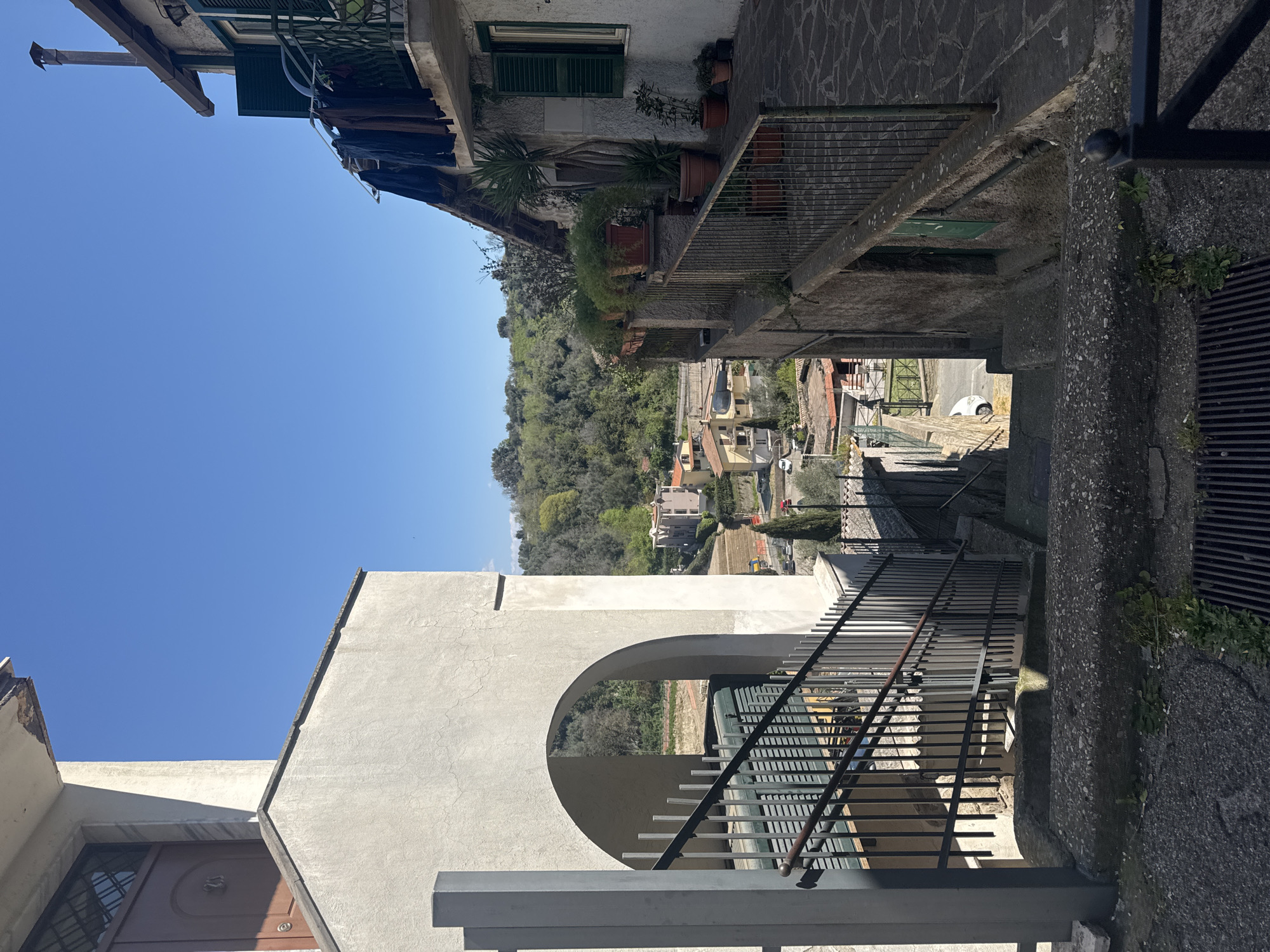 The image captures a narrow street scene in Castelnuovo di Porto, Italy, taken on a sunny day. The view is from an elevated position looking down an incline. The street is flanked by buildings with varied architectural details, including balconies with potted plants, stone facades, and aged walls. The sky is clear blue. A prominent feature is a modern archway structure in the foreground, with a metal railing. Below, the scene extends to show houses and greenery, characteristic of the town's landscape. A few cars are visible parked on the street below. The overall impression is a blend of historical architecture and contemporary elements set within the urban environment.