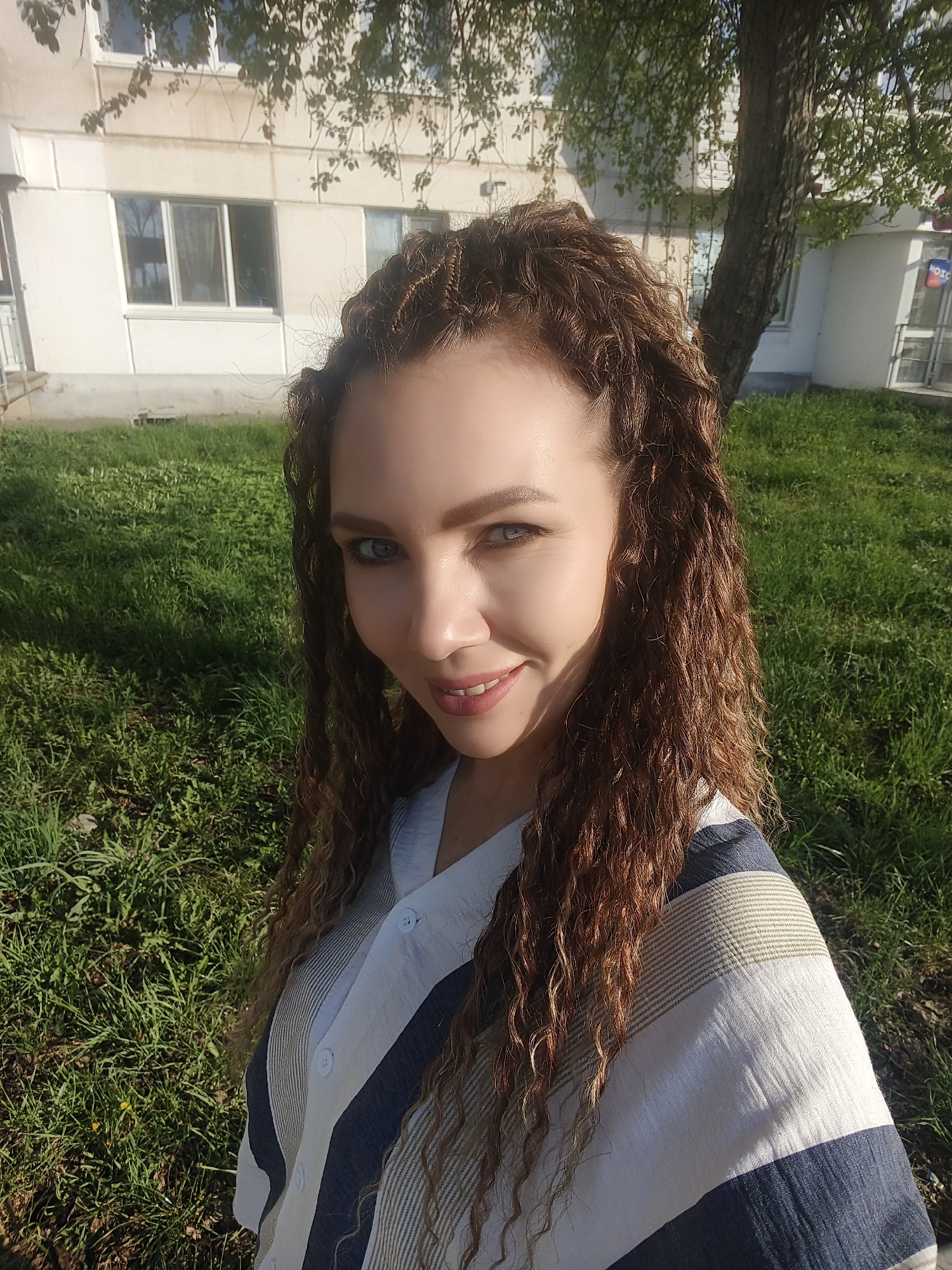 A medium shot depicts a woman with light blue eyes and long, curly brown hair, wearing a blue and white striped shirt with buttons. The woman smiles slightly at the viewer. The background shows a grassy lawn and a building with windows and what appears to be a tree. The location is Unknown, Unknown.