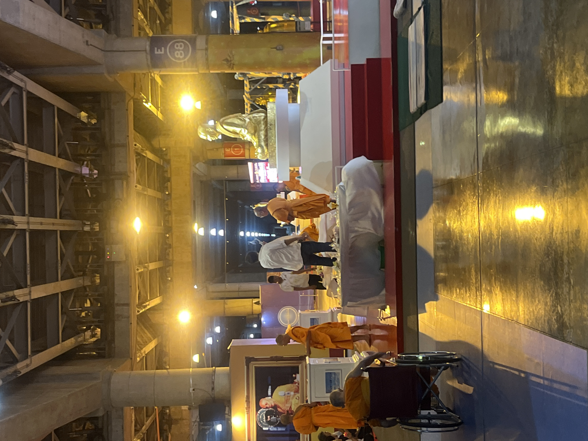 In a large, indoor, dimly lit space, possibly a covered market or transportation hub with exposed industrial-style ceilings and pillars, a ceremony appears to be underway. A golden statue of a seated figure, likely a religious icon, is centrally placed on a raised white platform with red steps leading up to it. Around the platform, several individuals in saffron robes, presumably monks, are gathered. One monk stands facing the statue, holding an object. Another monk is seated in a wheelchair in the foreground, facing away from the camera. Other individuals in casual clothing are also present, observing or participating. The floor is highly reflective, showing a golden sheen from overhead lighting. In the background, a large framed portrait of a person, also in saffron robes, is visible. A sign with the letter "E" and the number "88" is attached to one of the concrete pillars. The overall atmosphere suggests a formal or spiritual event.