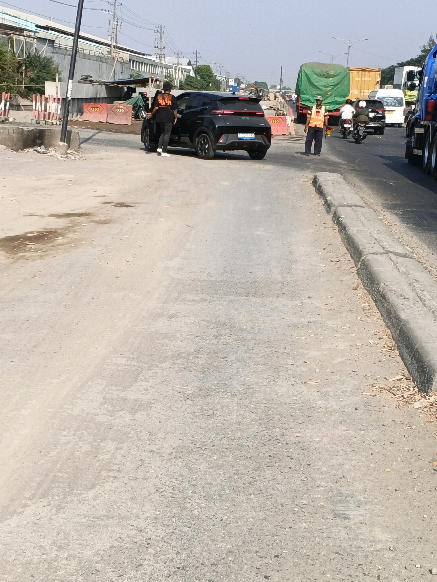 In Semarang, Indonesia, a black SUV is parked on a dusty, unfinished road, its passenger door ajar. Two individuals, one in a black t-shirt and jeans, the other partially obscured, stand near the vehicle. Beyond them, a traffic director in a bright orange vest stands in the middle of the road, overseeing a lane of traffic. Trucks, including a large green-covered vehicle and a blue semi-truck, are visible in the background, suggesting a busy transportation artery or construction zone. The environment is characterized by construction barriers, incomplete infrastructure, and a bright, sunny sky, indicative of an active construction site on a roadway in Semarang.