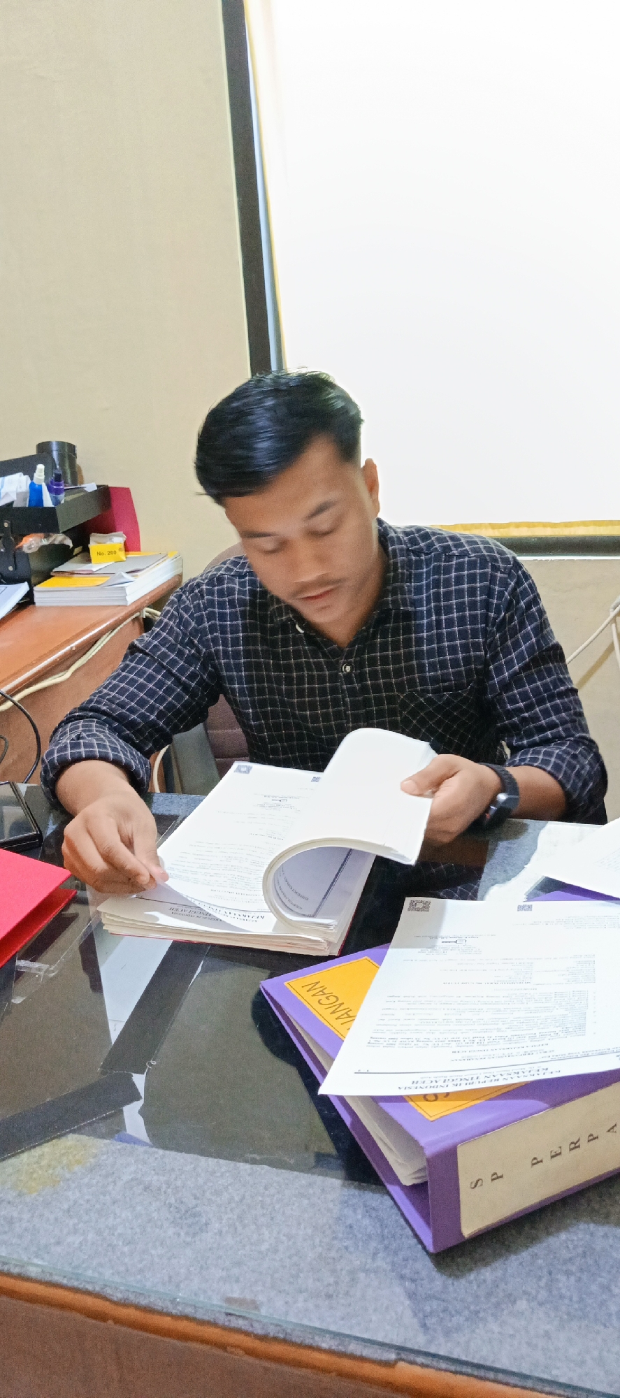 A young man with short, dark hair and a checked long-sleeved shirt is seated at a desk, looking down at a thick document he is holding open with his hands. The document has a white cover and appears to be a book or a large report. His left hand is holding the pages open while his right hand is positioned near the spine. The desk is made of dark, reflective material and is cluttered with various items, including folders, papers, and a red folder to the left. A stack of books or documents is visible on the left side of the desk, behind the red folder. A black object, possibly a scanner or printer, is also present on the desk. The background shows a light-colored wall and a white surface, possibly a screen or a window. The scene suggests an indoor office or study environment. The presence of Indonesian text on the documents, such as "KEJAKSAAN REPUBLIK INDONESIA" visible on one of the folders, strongly indicates the location is in Indonesia, and more specifically, the context of legal or administrative documents suggests Banda Aceh, given the prevalence of such documents in official settings within the city.