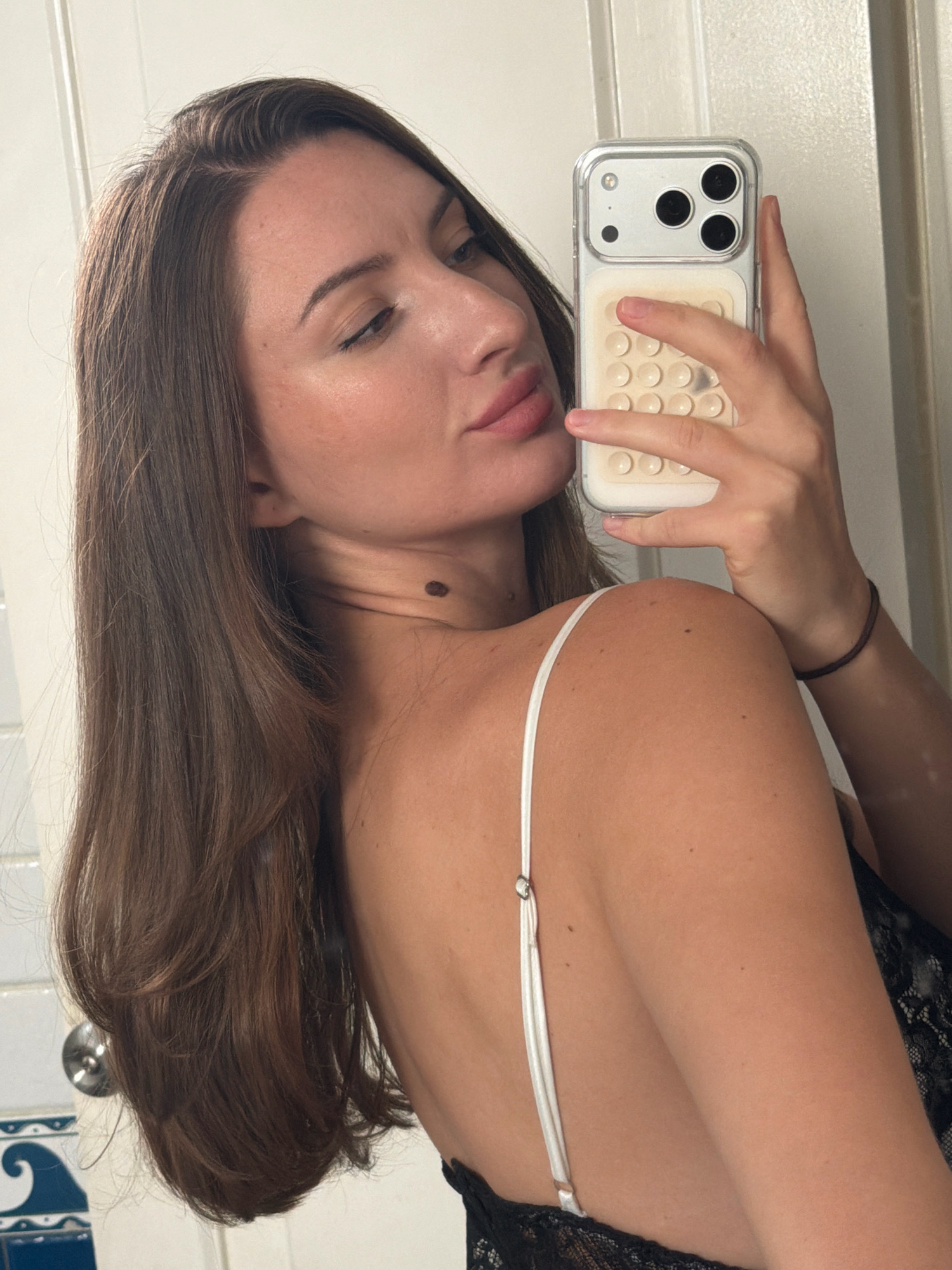 A woman with long, dark brown hair is taking a selfie in a dimly lit room. She is wearing a black lace top and a white strap from an undergarment is visible. Her skin is fair and she has makeup on. She is holding a smartphone in her right hand. The background appears to be a bathroom with white tiled walls and a mirror. There is no indication of the location being Sharm El Sheikh, Egypt.