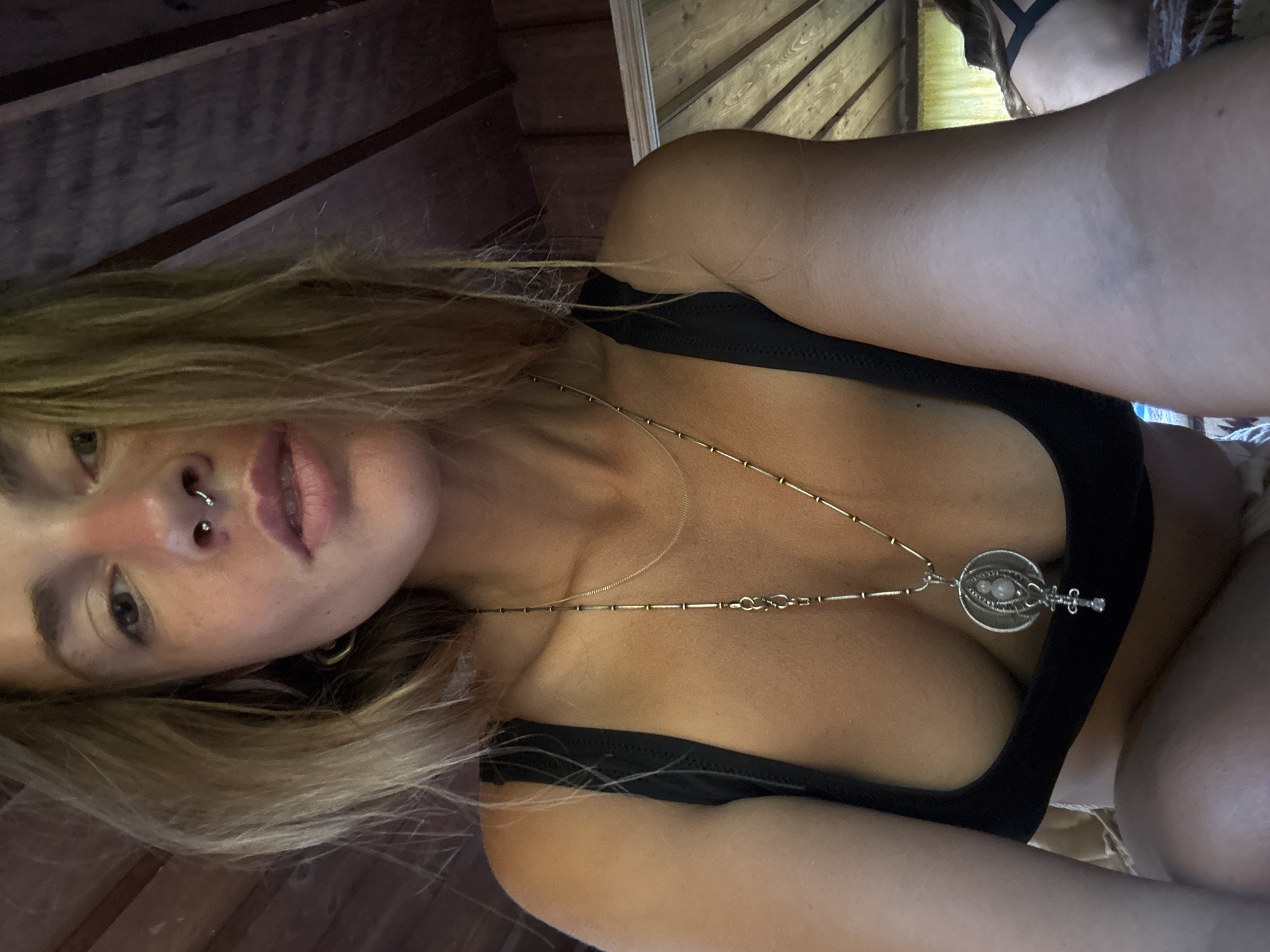 A close-up, low-angle selfie shows a woman with blonde hair and fair skin, looking directly at the camera. She has a septum piercing and her mouth is slightly open. Her eyes are brown, and there's a hint of blush on her cheeks. She is wearing a black bikini top and a long, silver necklace with a circular pendant. The background appears to be a rustic wooden interior, possibly a cabin, with exposed beams on the ceiling. Part of another person, wearing a similar bikini top, is visible in the background on the right side of the frame. The lighting suggests an indoor setting with natural light coming from the side.