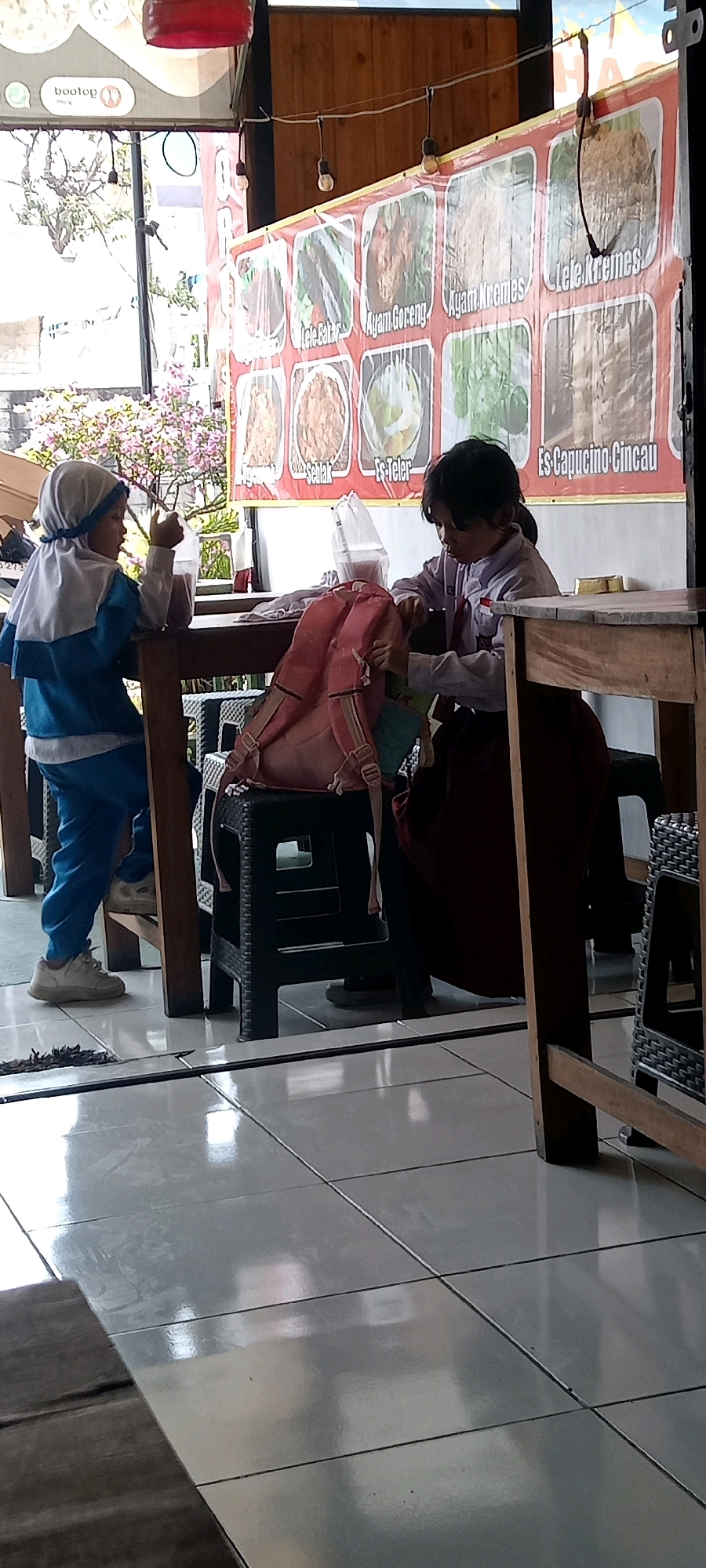 In the city of Pondok, Indonesia, a scene unfolds inside a restaurant, likely a food stall or cafe, with a banner displaying various menu items. Two young girls, dressed in school uniforms, are seated at a wooden table. The girl on the left, wearing a blue and white uniform, has a light blue hijab and is holding a transparent plastic cup with a reddish liquid. The girl on the right, in a white shirt and dark skirt, is engaged with a pink backpack on a black plastic stool. The backpack is open, and she appears to be looking inside it. The floor is tiled with a reflective surface. The environment suggests a casual dining area, with exposed wooden beams overhead and string lights. The overall lighting is subdued, with natural light entering from a window or opening in the background, illuminating some of the flowering plants outside.