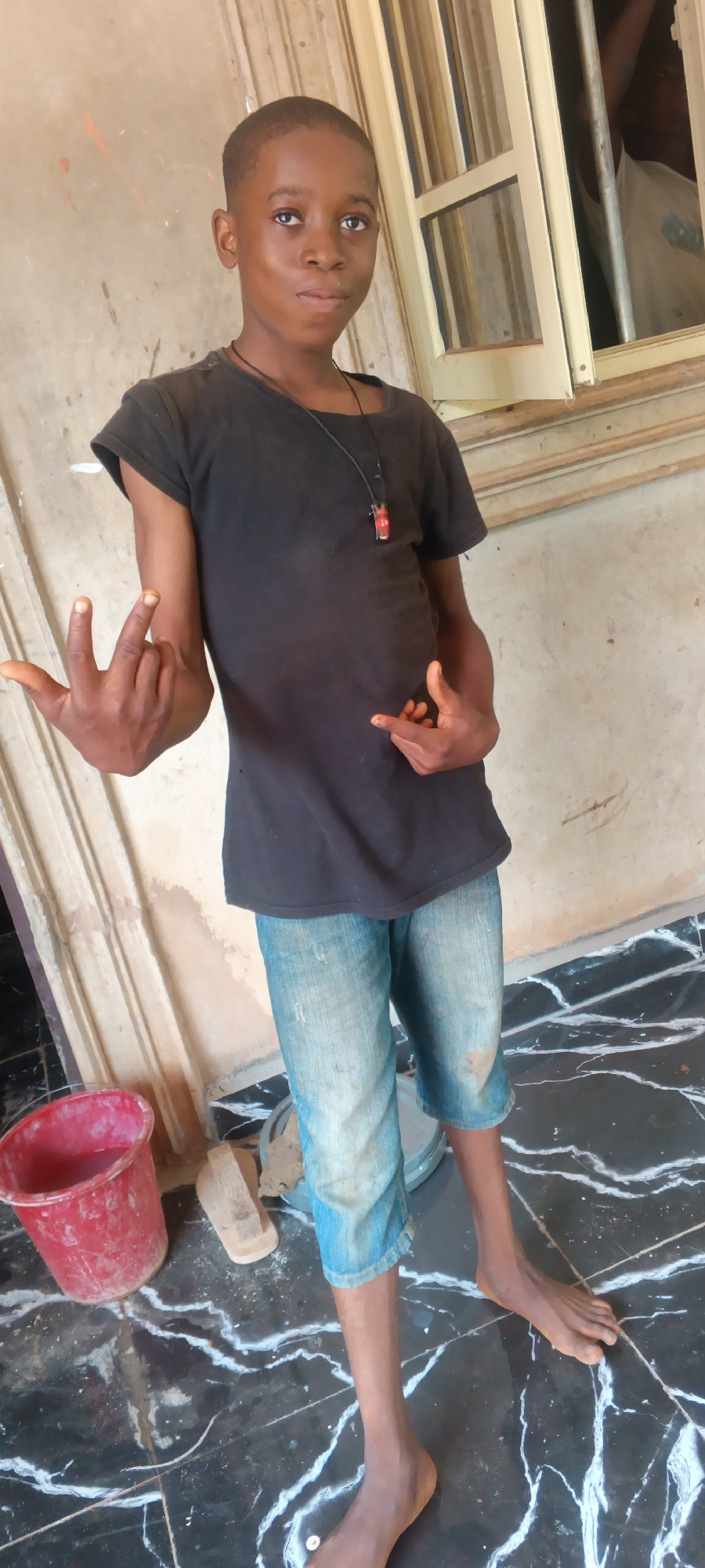 A barefoot, dark-skinned youth stands on a black and white marble-patterned floor. He wears a black t-shirt, and blue, knee-length denim pants. He makes gestures with both hands. A red bucket, a wooden plank and a metal container sit near his feet. He is standing in front of a wall, close to a window. Based on the context, the image was taken in Eziudo, Nigeria.