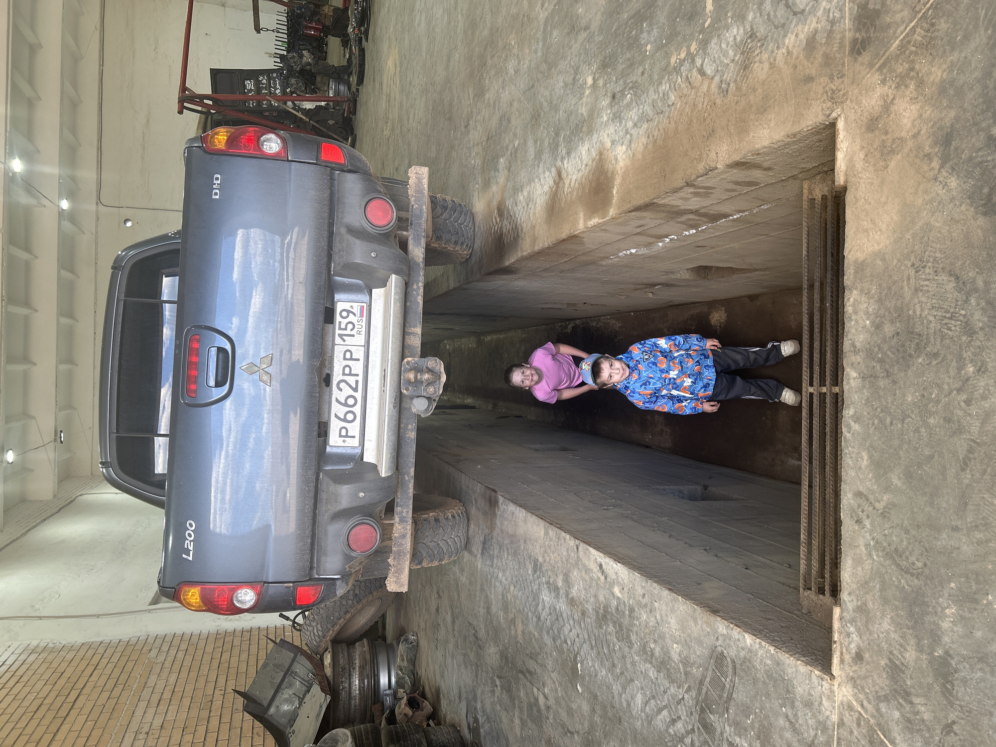 The image shows a gray Mitsubishi L200 pickup truck parked over a maintenance pit in what appears to be a garage or workshop. A person in a pink shirt and another person in a blue patterned jacket lie in the pit. The truck's license plate reads "P662PP 159 RUS". The environment is a workshop with equipment in the background and old tires and scrap metal to the side of the vehicle. Based on the license plate, this scene is likely in Russia. The presence of an L200 model in Chaikovsky, Russia, indicates the location is likely within this city.