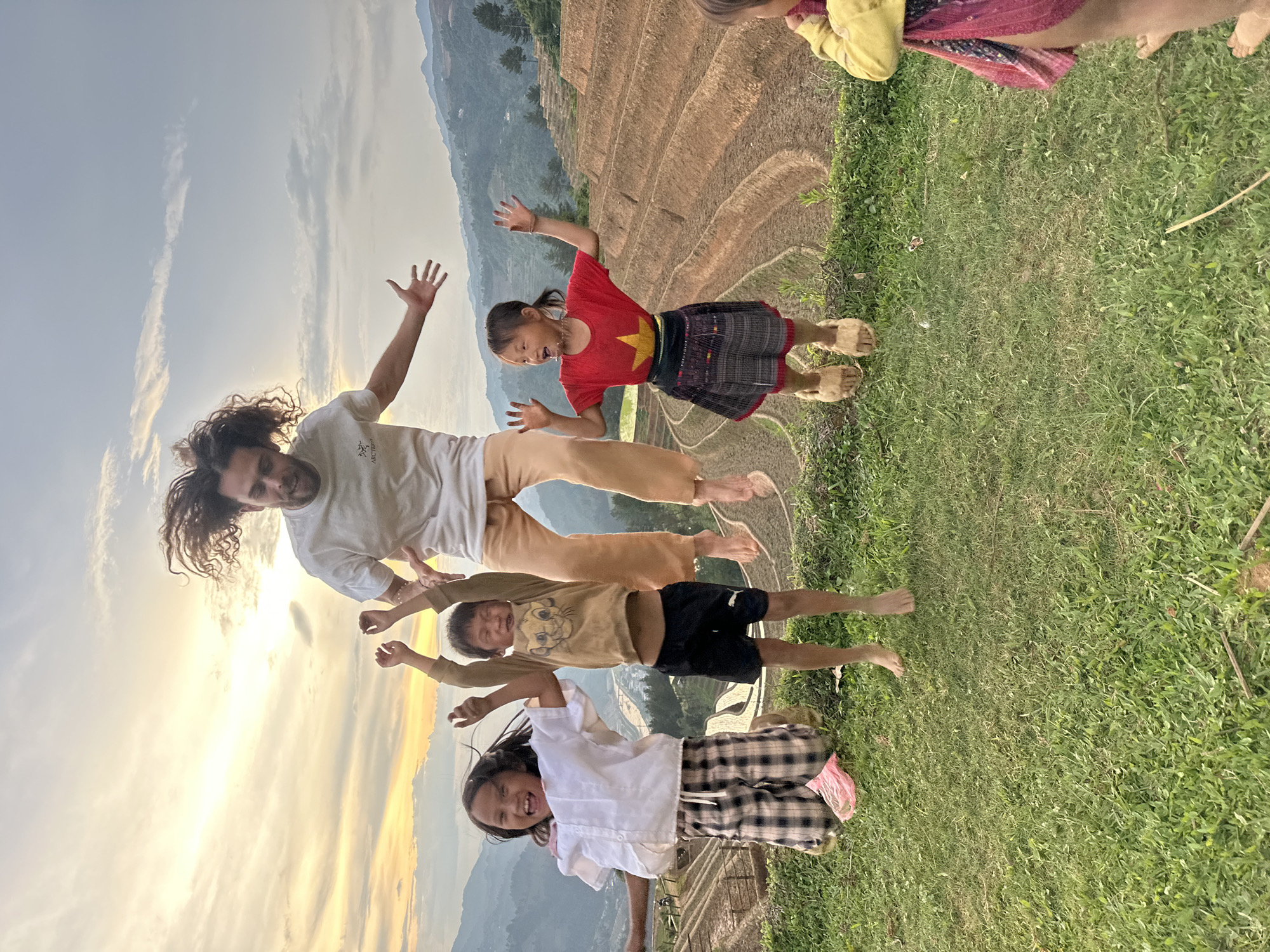 The image captures a lively moment with a man and four children joyfully jumping outdoors in a scenic, rural landscape. All five individuals are caught mid-air, with arms raised and expressions of excitement or delight.
The setting features expansive terraced rice fields, indicative of the Mù Cang Chải region in Vietnam, stretching across the background and climbing distant hills. The sky is bright with a low sun, suggesting late afternoon or early evening, casting a golden glow and creating a halo effect around the man's curly hair. The foreground consists of vibrant green grass where the group is jumping.
The man wears a white t-shirt, on which a faint "ARC'TERYX" logo is visible, and light-colored pants. The children are dressed in various casual attire: one girl in a white shirt and plaid pants, a boy in a tan animal-print t-shirt and dark shorts, and another girl in a red t-shirt with a yellow star (resembling the Vietnamese flag), a patterned dark skirt, and light-colored sandals. Parts of additional children are visible on the far right. The overall atmosphere is one of shared happiness and playful energy amidst a beautiful natural environment.