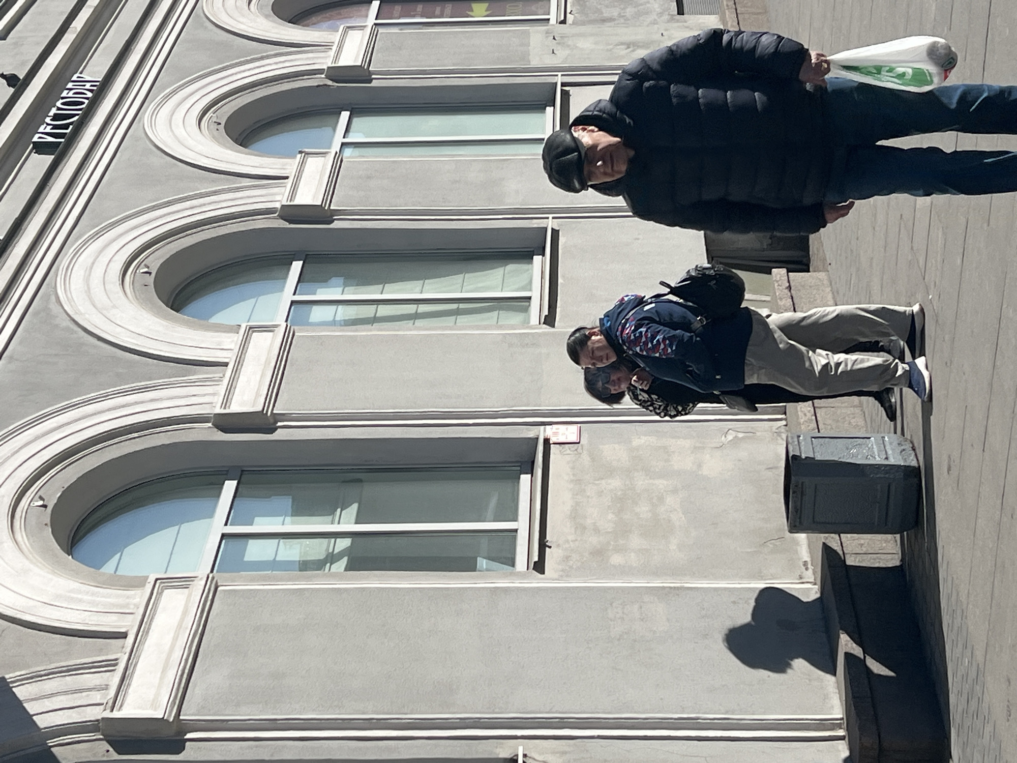 The image depicts a street scene in Saint Petersburg, Russia, on a bright, sunny day. The dominant feature is a multi-story building with a light grey facade on the left. The architecture is notable for its numerous arched windows, each framed by decorative light-colored moldings, giving it a classic or ornate appearance. Horizontal moldings further delineate the floors of the building. A sign with the Cyrillic text "РЕСТОРАН" (RESTORAN, meaning restaurant) is visible vertically mounted on the building's upper facade.
On the paved sidewalk or street to the right of the building, two distinct individuals and a partially visible third person are present. In the foreground, a man wearing a dark puffer jacket, dark pants, and a dark cap is walking towards the right side of the frame. He is carrying a white plastic bag with green text in his right hand. His strong shadow is cast on the ground to his right, indicating a clear, sunny day, likely around midday. Further back and to his left, another individual is visible, wearing a dark jacket with colorful patterns on the shoulders and light-colored pants, possibly carrying a backpack. A third person's head is partly visible behind this second individual. A dark-colored trash can stands on the sidewalk near the second person. The scene conveys a casual, daytime urban environment.