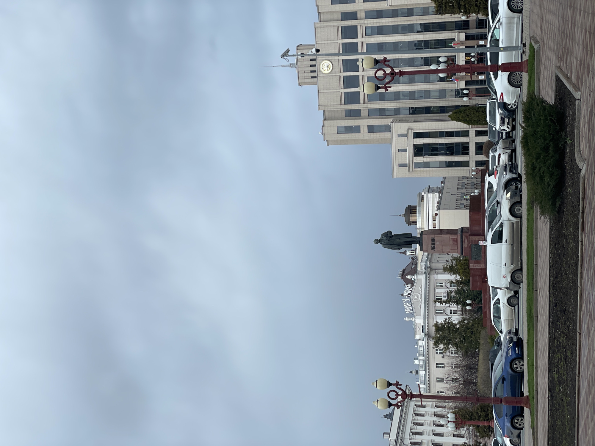 This image depicts an urban street scene in Kazan, Russia, under an overcast sky. The perspective showcases a blend of historical and modern architecture.
On the left side of the frame, a dark-colored statue of Vladimir Lenin stands atop a pedestal, clearly labeled with the Cyrillic text "ЛЕНИН" (LENIN). Below and behind the statue, an older, light-colored building is visible, featuring the prominent Cyrillic inscription "МИР'У - МИР" (Peace to the world) on its facade.
To the right, a tall, contemporary multi-story building dominates the skyline. It features a modern facade of glass and light-toned panels, with a distinctive round clock face near its upper floors. In front of this building, a row of parked cars, predominantly white, lines the street. Ornate, dark-colored lampposts with multiple light fixtures are spaced along the sidewalk, which is bordered by neatly maintained hedges and grassy areas.
The street itself is filled with various parked vehicles, including sedans and vans, indicating a busy urban environment. The sky is uniformly grey and cloudy, suggesting an overcast day without direct sunlight. There are no people visible, and no specific event or activity is taking place, presenting a static, everyday snapshot of the city. The presence of the Lenin statue and the visible Cyrillic text reinforce the cultural and geographical context of Kazan.