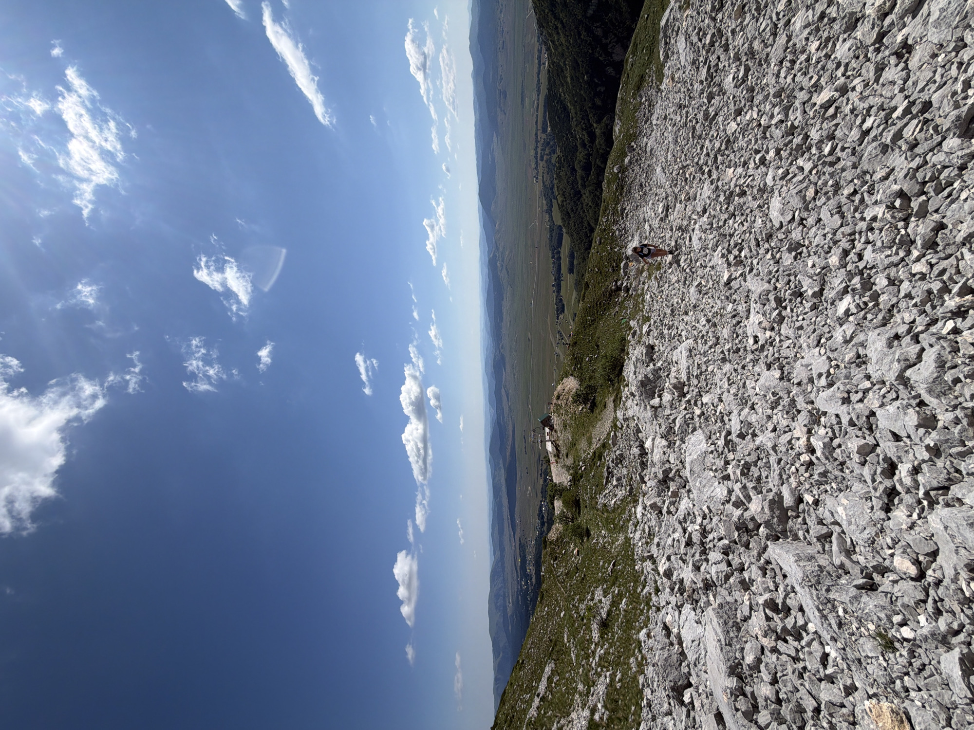 An aerial, wide-angle shot depicts a person hiking down a rocky, mountainous terrain with a vast valley stretching out in the distance under a clear blue sky dotted with white clouds. The foreground is dominated by grey rocks and sparse green vegetation, with a single individual, wearing dark shorts and a t-shirt, walking downhill on a path strewn with smaller rocks. The mid-ground reveals more green slopes, a small structure with a red roof and a white fence, and a ski lift. The background showcases rolling hills and mountains under a bright, sunlit sky. The scene is located near Pašina voda in Montenegro.