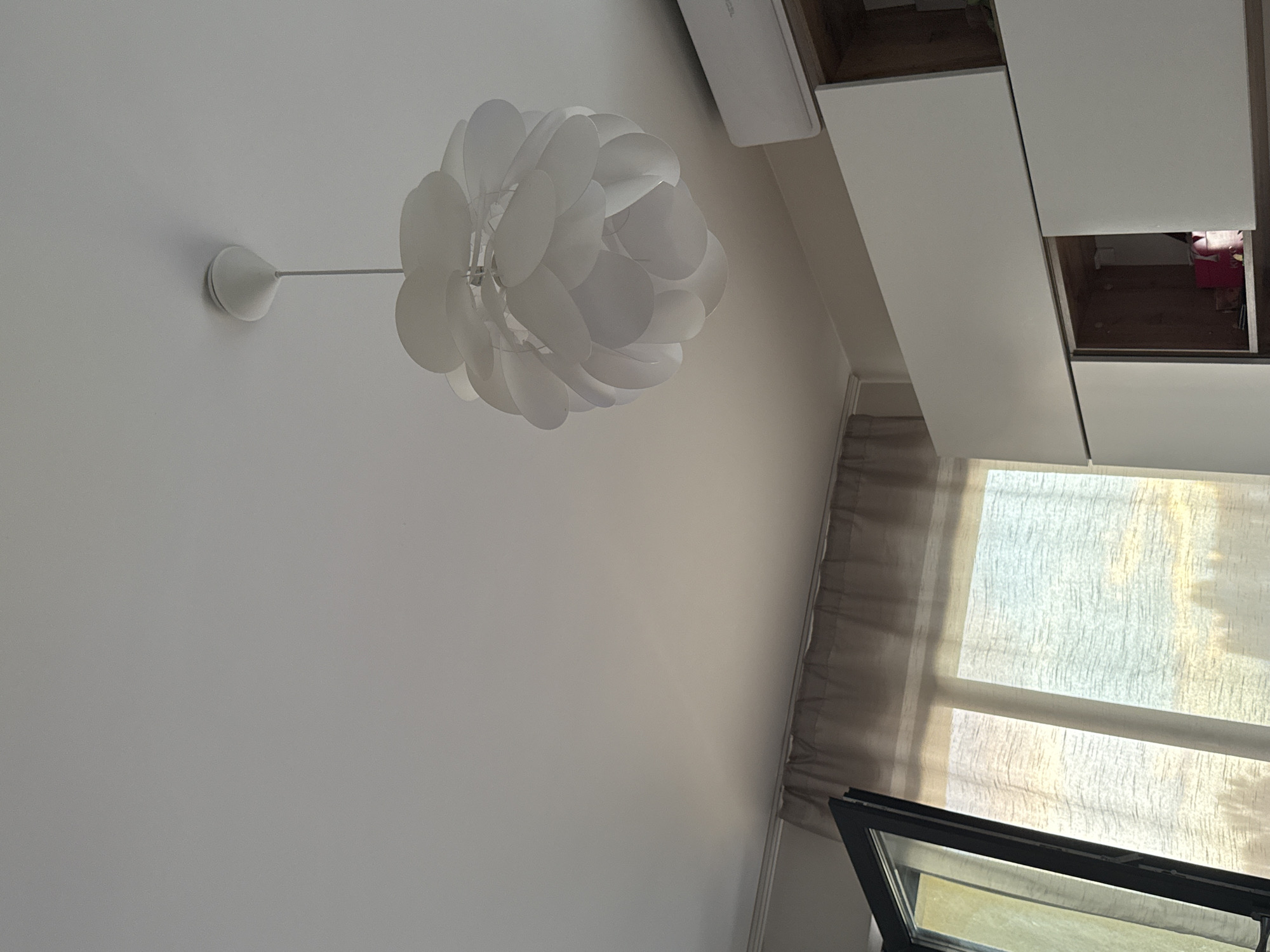 The image depicts an indoor scene with a white ceiling. A modern, spherical pendant lamp with translucent white petal-like diffusers hangs from the ceiling. To the right, a white air conditioning unit is mounted on the wall. Below the air conditioner, a section of white cabinetry with a wooden interior is visible. On the left side of the image, a window is partially open, revealing sheer curtains that allow natural light to filter through, illuminating the scene with a soft glow. The curtains appear to be a light beige or taupe color. The overall ambiance is bright and airy. No specific location in Sofia, Bulgaria, can be identified from the visual information provided.