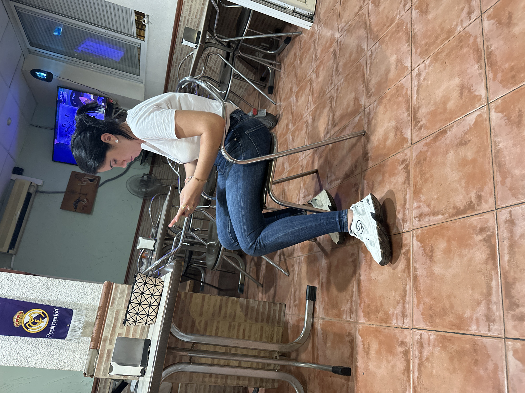 The image depicts an indoor scene, likely a cafe or restaurant in Alicante, Spain. A woman with dark hair pulled back in a ponytail is seated at a table, looking down at a tablet she is holding. She is wearing a white t-shirt, blue jeans, and white athletic shoes. The table has a metallic frame and a tiled top. On the table are a napkin dispenser and a small, patterned pouch. In the background, there are other tables and chairs, and a television screen displaying content. A Real Madrid banner hangs on the wall to the left. The floor is tiled with reddish-brown square tiles. The lighting appears to be artificial.