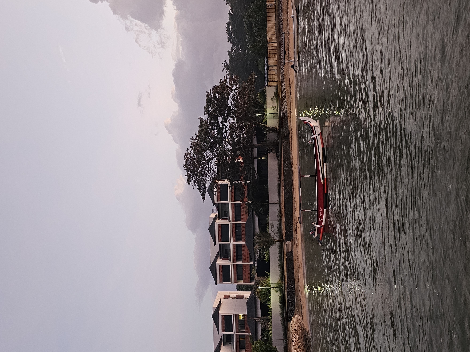 The image depicts a serene waterside scene at what appears to be a resort or hotel in Denpasar, Indonesia, likely during the late afternoon or early evening. In the foreground, calm, dark water reflects the soft light of the dusky sky, with subtle ripples breaking the surface. A traditional Indonesian outrigger boat, painted red and white with black trim, is moored in the water, its reflection shimmering on the surface. Another, smaller white boat with an outboard motor is visible further out.
The midground features a sandy beach lined with a low stone wall and a neatly maintained lawn. Behind the beach, a row of modern, multi-story buildings with white facades and dark-tiled roofs are prominent. These structures are architecturally similar, suggesting they are part of a cohesive complex. Large trees, one particularly broad and leafy, provide a natural element, casting shadows and adding to the tropical ambiance. The sky above is a soft gradient of pale purple and pink, with a few wispy clouds adding texture, indicative of twilight. The overall impression is one of tranquility and modern comfort set against a natural coastal backdrop.