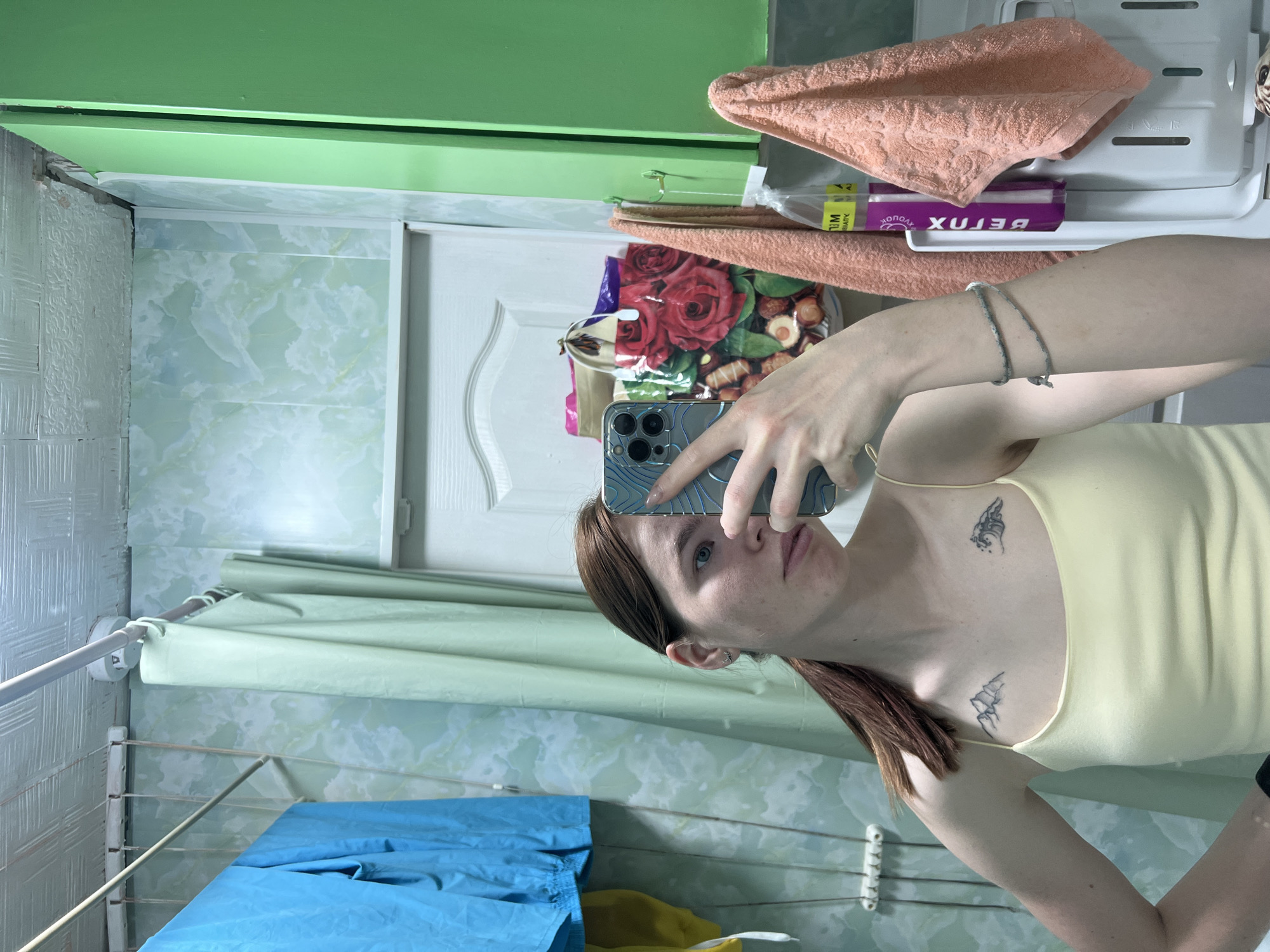 A woman is taking a selfie in a bathroom. The bathroom has light green marble patterned walls. Above a white door is a green painted section of the wall. Above the door, there is a shelf with an orange towel, a package of "Betox" beauty supplements, and a bag with a floral pattern. To the left of the woman is a shower curtain on a metal rod above a drying rack with blue and yellow clothes hanging from it. The woman has red hair and is wearing a yellow tank top. The woman has two tattoos on her chest. She is holding a blue iPhone. The location is the city of Chaikovsky, Russia.
