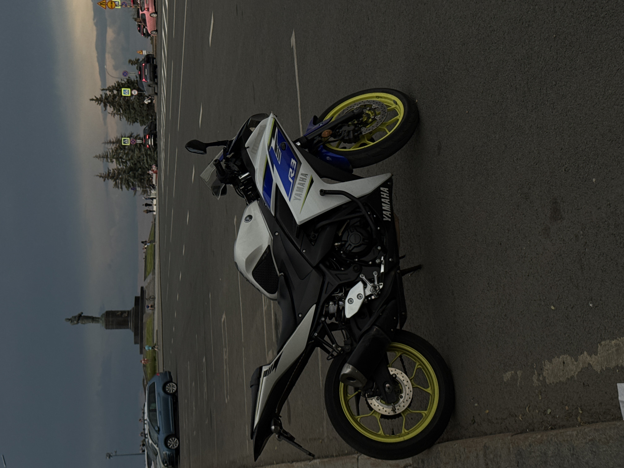 A Yamaha R3 motorcycle is parked on the side of a street in an urban environment. The motorcycle is white with blue and black accents, and it has bright yellow rims. It is positioned facing towards the right side of the frame, with its front wheel slightly turned. The motorcycle is parked on asphalt, next to a curb. In the background, a statue on a pedestal stands prominently, with a few trees and people visible near it. Further in the background, cars are seen on a road, and the sky is overcast with dark clouds. The overall scene suggests a daytime setting under a cloudy sky.