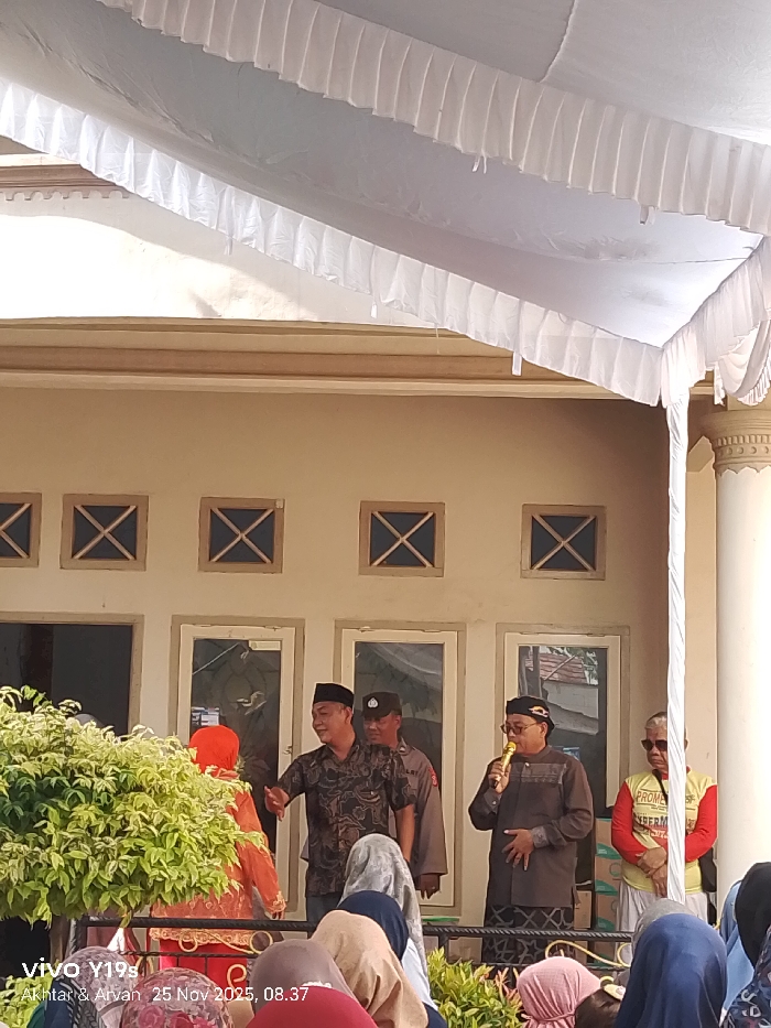 A group of people are gathered under a white tent, in front of a light-colored building with symmetrically placed windows. Three men stand behind a short barrier, two are wearing caps, and one is speaking into a microphone. A uniformed officer is also present. In the foreground, the heads and shoulders of the audience are visible. The image has a time and date stamp in the lower left corner.