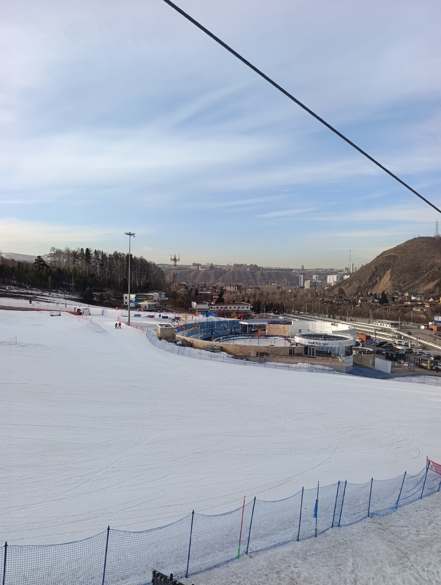 The image shows a ski slope at the Bobrovy Log Fun Park in Krasnoyarsk, Russia. The slope is covered in snow and is mostly flat in the foreground. A blue safety net runs along the lower edge of the frame. In the middle ground, there are buildings and facilities associated with the park, including Hakka-ski rental. Behind these buildings is a road with cars parked along it. Beyond the buildings, the city of Krasnoyarsk is visible, with apartment buildings on the far side. A large hill is to the right of the city. Two people are seen skiing. A ski lift cable is visible in the upper right corner. The sky is blue with streaks of white clouds.