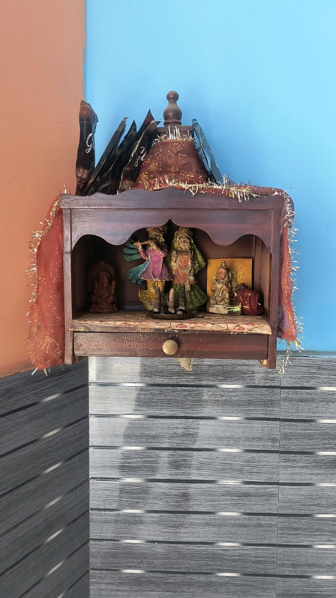 The image shows a small wooden altar or shrine placed against a wall. The altar is dark brown and has two shelves. The top shelf has a decorative red cloth with gold trim, and several dark, folded items are placed behind it. The bottom shelf contains several religious figurines. On the left, there is a statue of Ganesha. In the center, there are two figurines of Radha and Krishna, with Krishna playing a flute. To the right of them, there is a smaller figurine of a seated deity. The altar has a drawer at the bottom with a round knob. The wall behind the altar is painted in two colors: orange on the left and blue on the right. The wall has a distinct corner where the two colors meet. Below the altar, the wall transitions to a horizontally-slatted surface, with the slats appearing to be made of wood or a similar material, painted in shades of gray and white. The lighting suggests it is daytime. The scene is likely a domestic religious space.