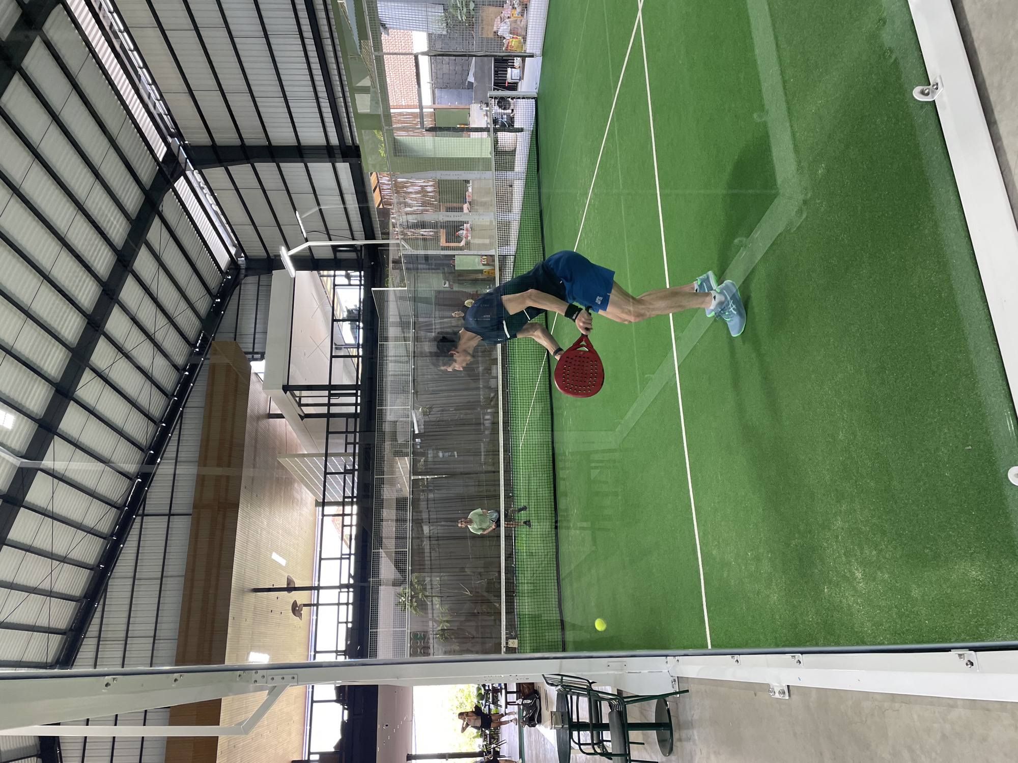 This image depicts an indoor padel tennis court in Kuta Selatan, Indonesia. The court has a green artificial turf surface with white lines marking the boundaries. A net divides the court in half.
In the foreground, a male player, wearing a dark t-shirt and blue shorts, is positioned on the right side of the court, holding a red padel racket. He is in the motion of playing, with his body turned and racket raised. His feet are clad in light blue and white athletic shoes. A yellow ball is visible in the air to his left.
Further back on the court, another male player, dressed in a green t-shirt and light shorts, is standing closer to the net on the left side, also appearing to be in the midst of a game.
In the background, beyond the court, a fence separates the playing area from a spectator or lounge area. Several tables and chairs are visible in this area, and two women are standing in the background, one in a black tank top and shorts, and the other in a white top and dark shorts.
The facility features a high, industrial-style roof with metal beams and translucent panels, allowing natural light to illuminate the court. Various light fixtures are also present. The walls of the facility appear to be constructed from metal and glass. Reflections are visible on the glass panels surrounding the court, creating a sense of depth and showing glimpses of the outdoor environment and other parts of the building.