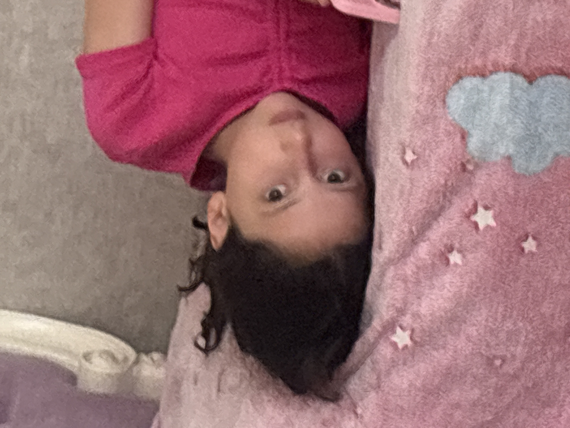 A young girl with dark, curly hair lies on a pink, star-patterned blanket, her head tilted towards the viewer. She wears a bright pink t-shirt. The background is a neutral gray, and to the left, a portion of a white decorative element of a bed is visible, possibly a headboard, with a purple soft surface behind it. The image is taken at a close-up perspective. The location in Georgiyevsk, Russia cannot be determined from the image alone.
