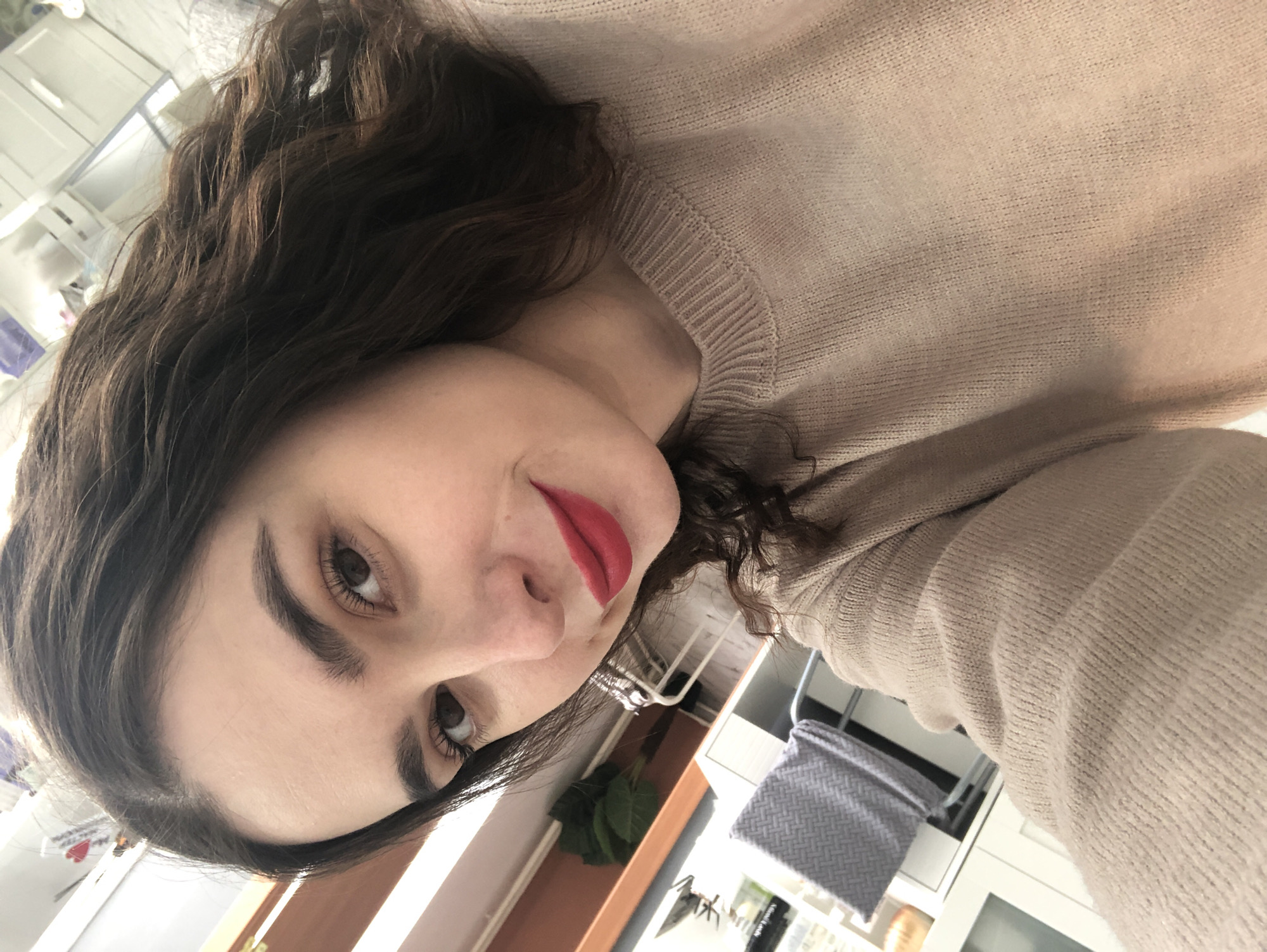 The image displays a close-up portrait of a woman taken indoors in Chaikovsky, Russia. The woman, with fair skin, features dark, neatly shaped eyebrows and dark eyes. She is wearing vibrant red lipstick, which is a prominent detail. Her dark brown hair is wavy or curly, flowing around her face and shoulders. She is dressed in a light beige or tan knitted sweater, showcasing a textured knit pattern, particularly noticeable around the collar. The woman looks directly towards the viewer with a calm expression, possibly a slight, subtle smile or smirk.
The background suggests a domestic setting. On the right side of the frame, there are white cabinets or shelves. Below them, a green leafy plant is visible, along with a grey textile item featuring a woven herringbone pattern, possibly a blanket or storage bin. The lighting appears natural, indicating it is likely daytime. There are no other distinct objects or activities captured, implying a simple portrait shot. No visible text is present in the image.