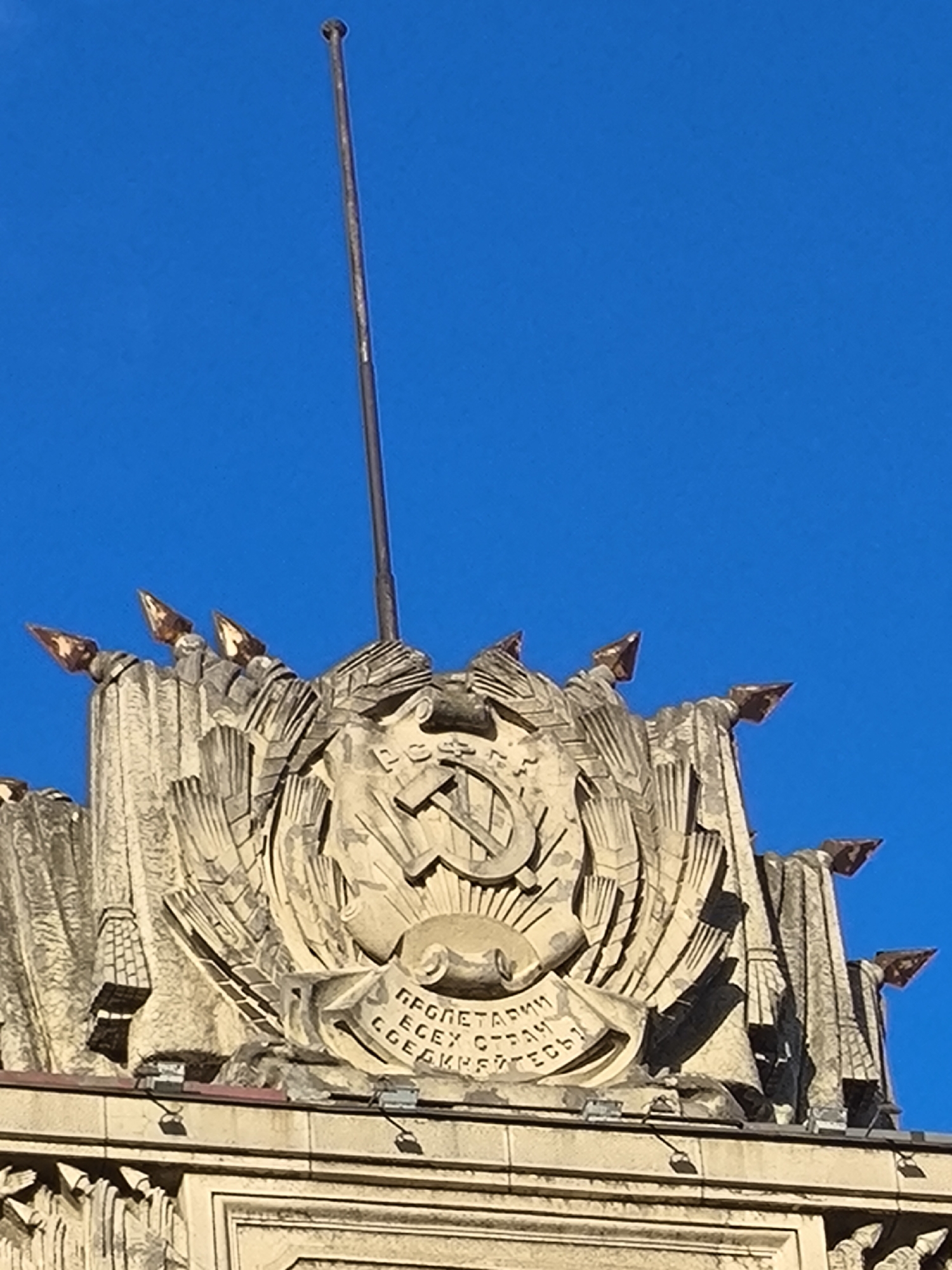 The image shows the top of a building against a clear blue sky. The building is adorned with a large, ornate stone relief. The relief features the hammer and sickle symbol, a prominent emblem of the Soviet Union, surrounded by stylized wheat sheaves and a rising sun. Below this, a banner is carved with Cyrillic text that reads "Пролетарии всех стран, соединяйтесь!" which translates to "Workers of the world, unite!". The architectural style appears to be Soviet Constructivism or Stalinist architecture, common in the mid-20th century.
The building's facade is decorated with sharp, angular elements and what appear to be metallic spearhead-like ornaments along the upper edge, catching the sunlight and appearing coppery or bronze. A slender metal flagpole stands vertically behind the central relief, extending upwards out of frame. The lighting suggests it is daytime, with the sun casting distinct shadows on the relief, highlighting its three-dimensional details. The overall scene conveys a sense of monumental grandeur and historical significance, typical of Soviet-era public buildings. The location is specified as Saint Petersburg, Russia, a city with numerous examples of such architecture.