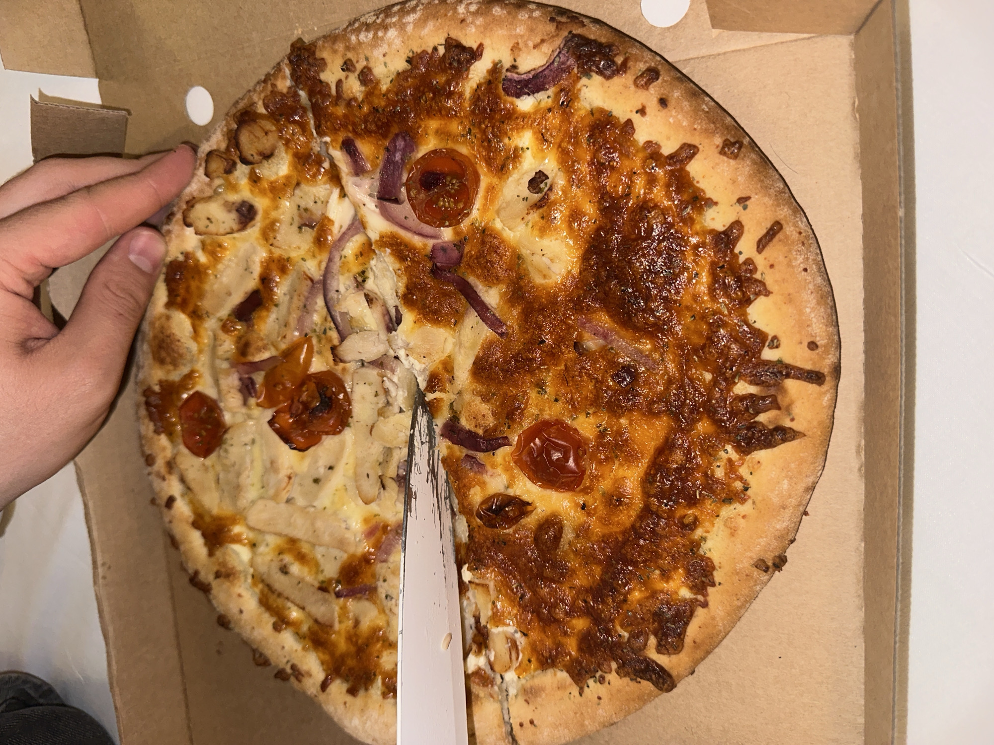 A photograph depicts a pizza in a cardboard box, situated in Ramonville-Saint-Agne, France. A fair-skinned hand is seen touching the edge of the pizza, while a large, silver knife divides the pizza in half. The pizza has a light-colored crust with toppings including tomatoes, red onions and strips of white food. One half is baked to a light golden brown, while the other half is a darker golden brown color. The pizza is sitting in a tan cardboard box. The surface under the box is white. The person wearing black pants can be seen to the lower left.