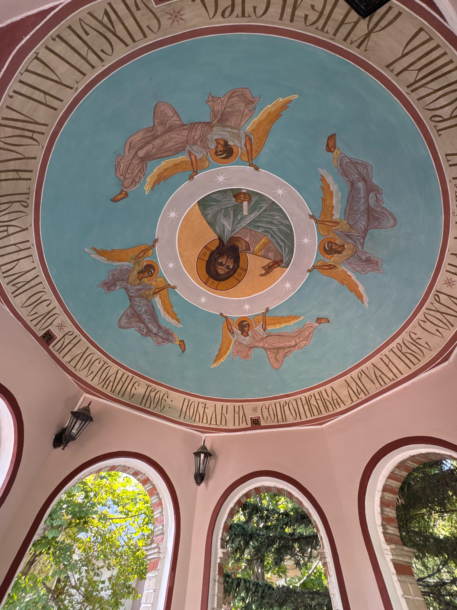 The image depicts a fresco on the ceiling of a domed structure, possibly a church or monastery, in Moscow, Russia. The fresco features a circular composition with religious imagery. At the center, a haloed figure resembling Jesus Christ is depicted, surrounded by four winged figures, presumably angels, in dynamic poses. These angels are rendered in muted tones of pink and blue, with golden wings. The entire scene is framed by a wide band containing Cyrillic inscriptions in a dark brown color, bordered by maroon and a lighter brown. Below the dome, the interior of the structure is visible, with arched openings revealing lush green trees and foliage outside. Two dark, ornate lanterns are mounted on the light pink walls of the structure, one on each side of the central archway. The lighting suggests it is daytime.