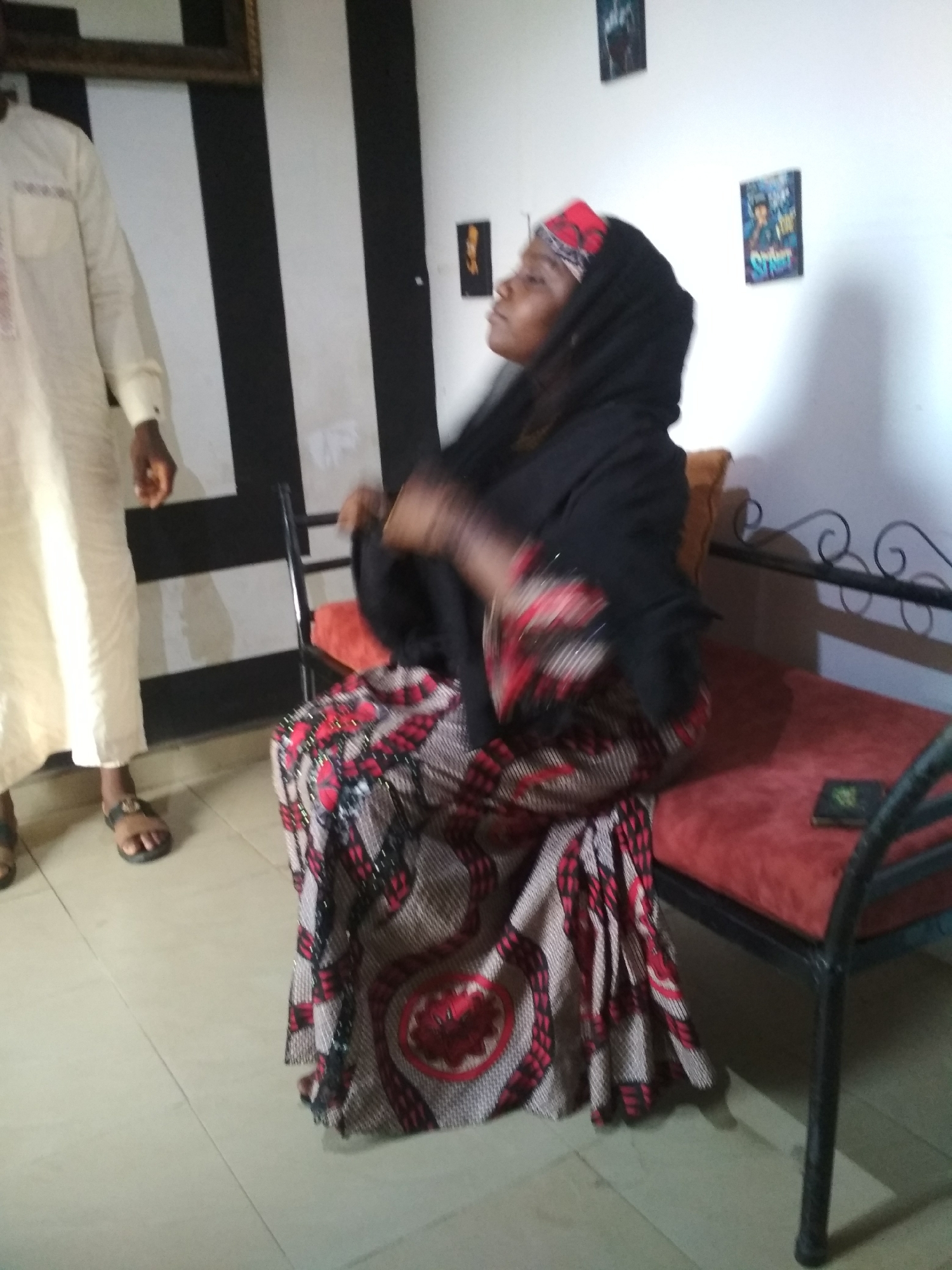 The image depicts a woman in Jos, Nigeria seated on a metal-framed bench with a red cushion. She is wearing a black headscarf and a patterned dress with red and black accents. A man in a light-colored robe stands to her left. The background features a black and white striped wall and a white wall adorned with small framed pictures. A small book is visible on the bench beside the woman.