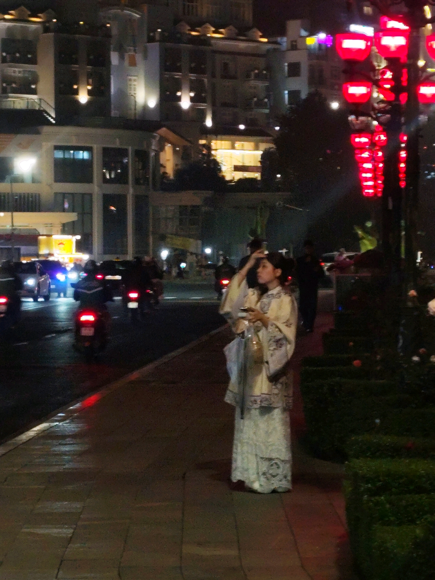 The image depicts a nighttime street scene in Da Lat, Vietnam. In the foreground, a woman wearing traditional Vietnamese attire, a yellow and white ao dai with intricate patterns, stands on a sidewalk. She is holding a phone and a bottle in her hands and her hair is partially covering her face as she looks upwards. The sidewalk is paved with brick-like stones, and the area is illuminated by ambient streetlights and the glow of red lanterns hanging from a pole on the right side of the image.
The background shows a busy street with vehicles, including cars and motorcycles, moving along. The buildings lining the street are brightly lit, indicating a commercial or residential area. The overall atmosphere is lively and illuminated by artificial lights. The presence of red lanterns suggests a festive occasion or a common decorative element in the area. The wet appearance of the sidewalk indicates recent rain or a damp environment.