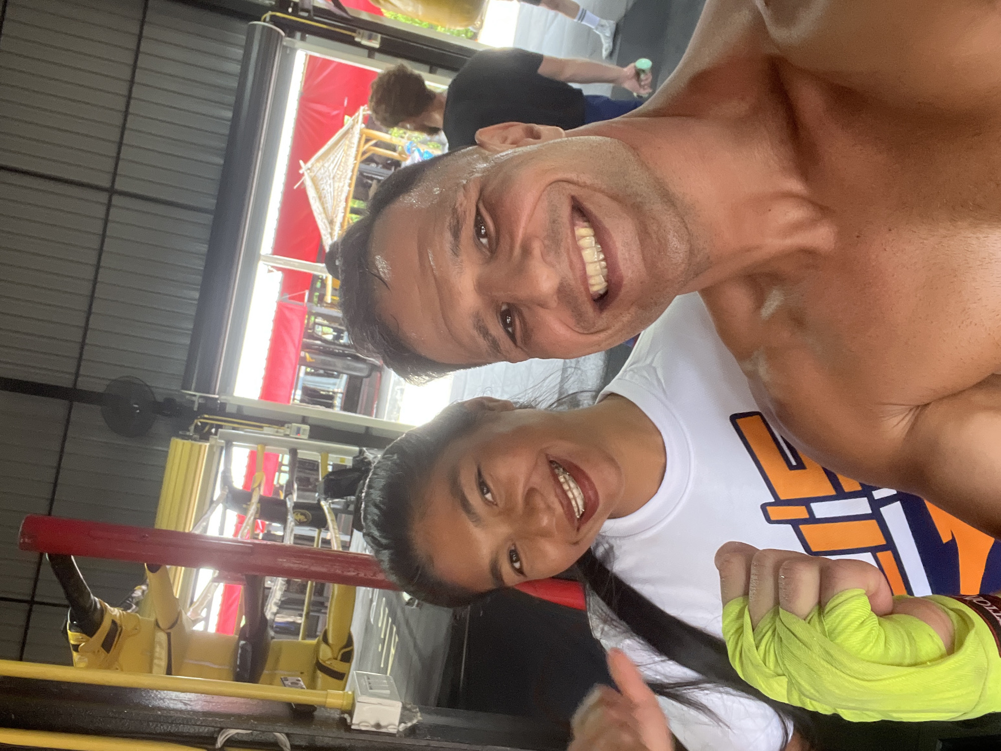 In Chalong, Thailand, a selfie is taken in a gym setting, possibly a boxing gym given the equipment visible in the background. The foreground is dominated by two individuals. A woman on the left, wearing a white t-shirt with an orange and blue graphic, smiles broadly, displaying braces. Her left hand, wrapped in bright yellow boxing hand wraps, is held up and slightly towards the camera. To her right, a man, shirtless and with sweat glistening on his skin, also smiles widely, his face filling a significant portion of the frame. In the background, a boxing ring with red ropes is visible, along with a punching bag and other gym apparatus. A person with curly hair wearing a black t-shirt is also seen in the mid-ground. The environment appears to be an open-air or partially open gym, with a corrugated metal roof overhead and red awnings visible in the distance.