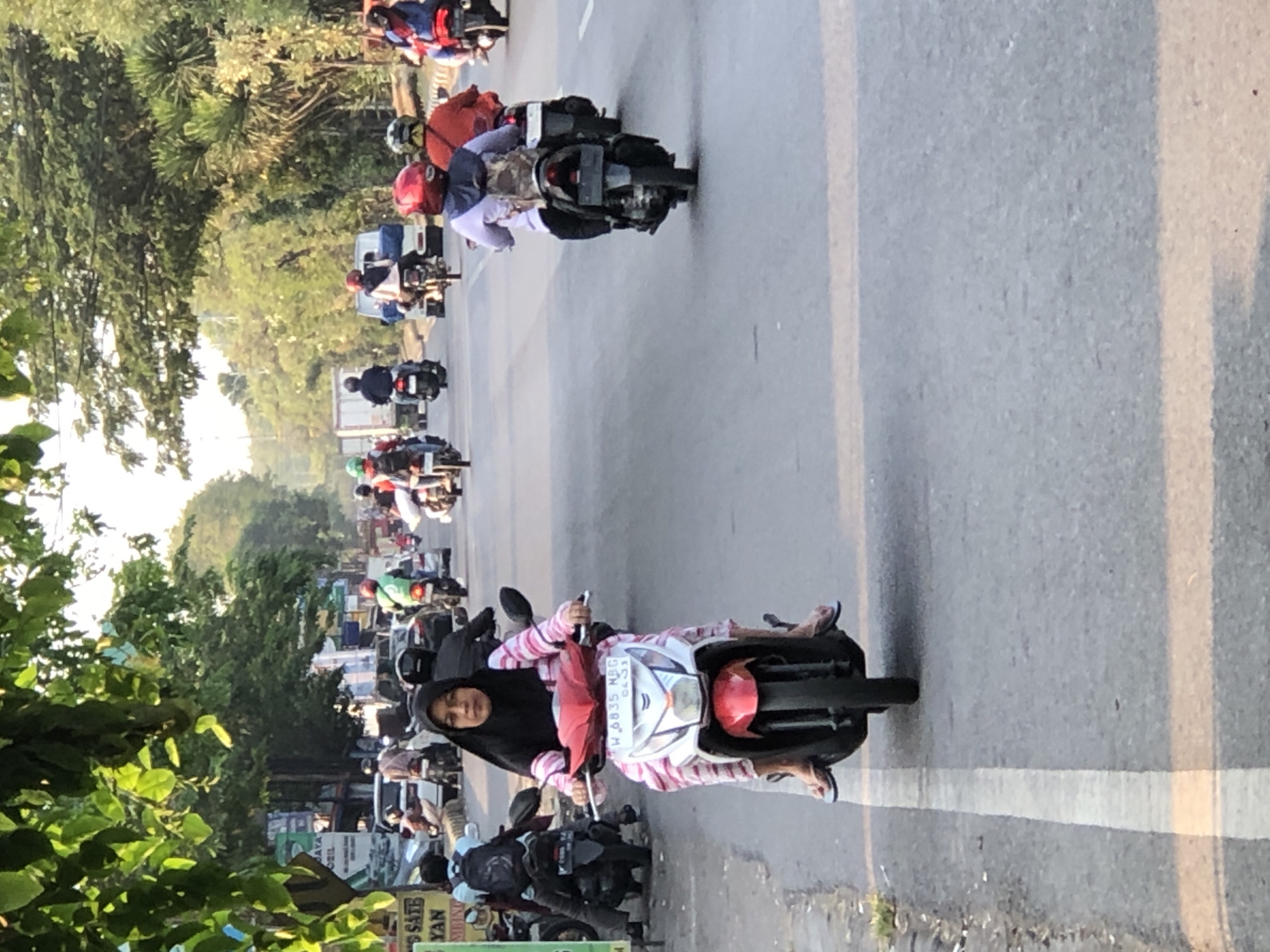 In Kludan, Indonesia, a street scene unfolds with multiple motorcycles in motion. The foreground features a woman wearing a black hijab and a pink and white striped shirt, riding a red and white motorcycle. She is wearing blue flip-flops. Behind her, other motorcycles are visible, some carrying passengers. The road is paved with distinct white and yellow lane markings. The surrounding environment includes trees and a glimpse of buildings and storefronts along the roadside. The lighting suggests it is late afternoon or early morning.