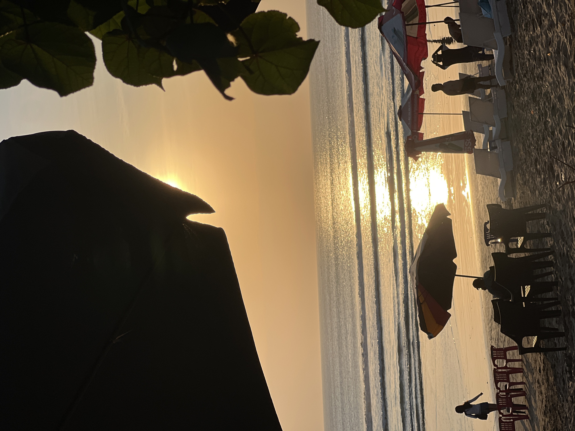 At sunset on Kuta Beach, Bali, Indonesia, the golden light reflects off the ocean, creating a shimmering path. The foreground is dominated by dark silhouettes of palm leaves and an umbrella casting shadows on the sand. Several beach chairs and umbrellas are scattered across the beach, some occupied by silhouetted figures. A lone woman walks along the shoreline, her figure small against the vastness of the ocean. The waves are gentle, rolling onto the sand in a rhythmic pattern. The sky is a soft gradient of orange and yellow, with a few wispy clouds.
