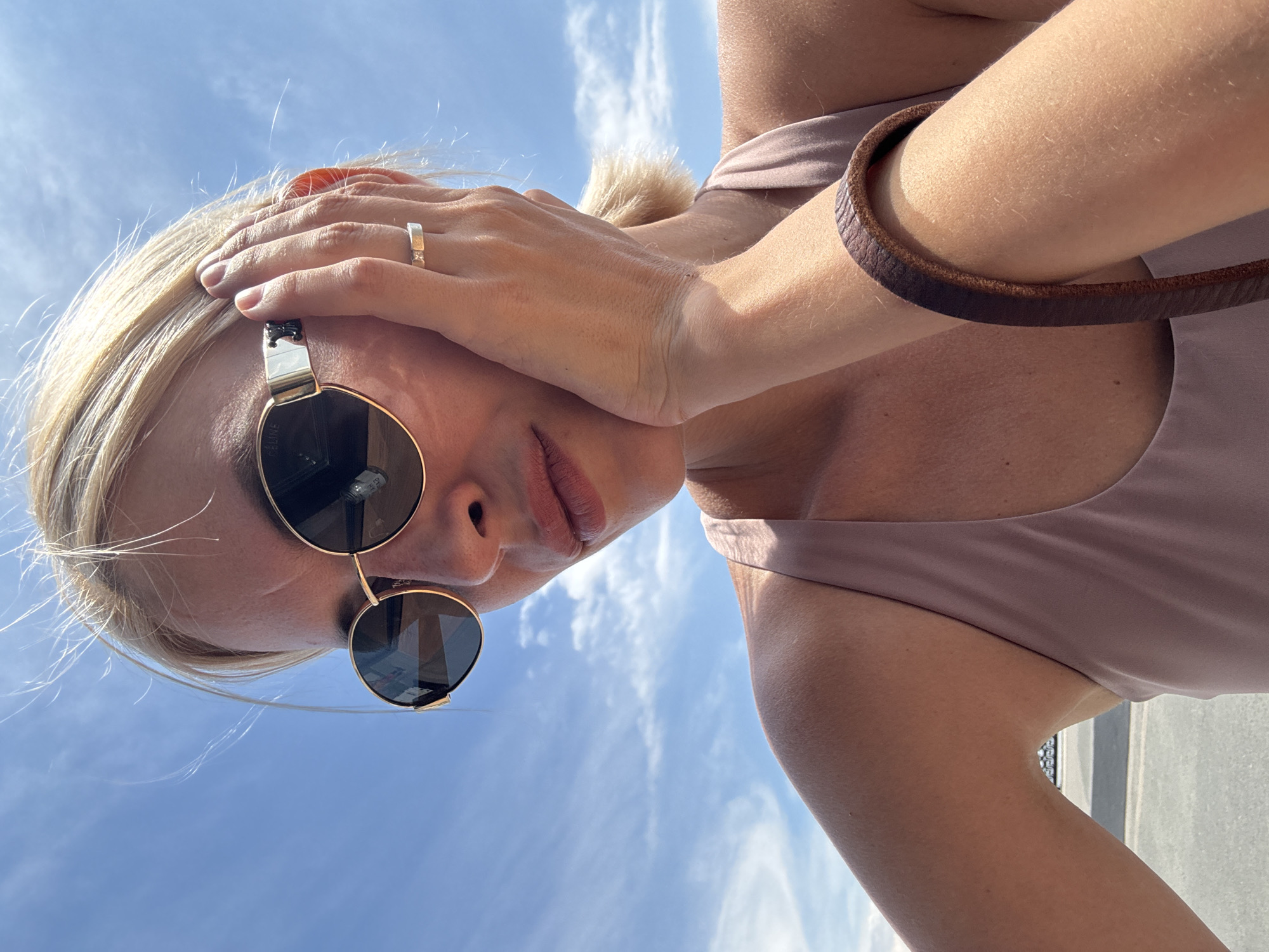A woman with blonde hair and sunglasses is captured in a close-up, low-angle shot against a bright blue sky with wispy clouds. She is wearing a light pink tank top and resting her hand on her cheek, adorned with a ring. A brown leather bracelet is visible on her wrist. The background appears to be an outdoor urban setting, possibly a parking lot.