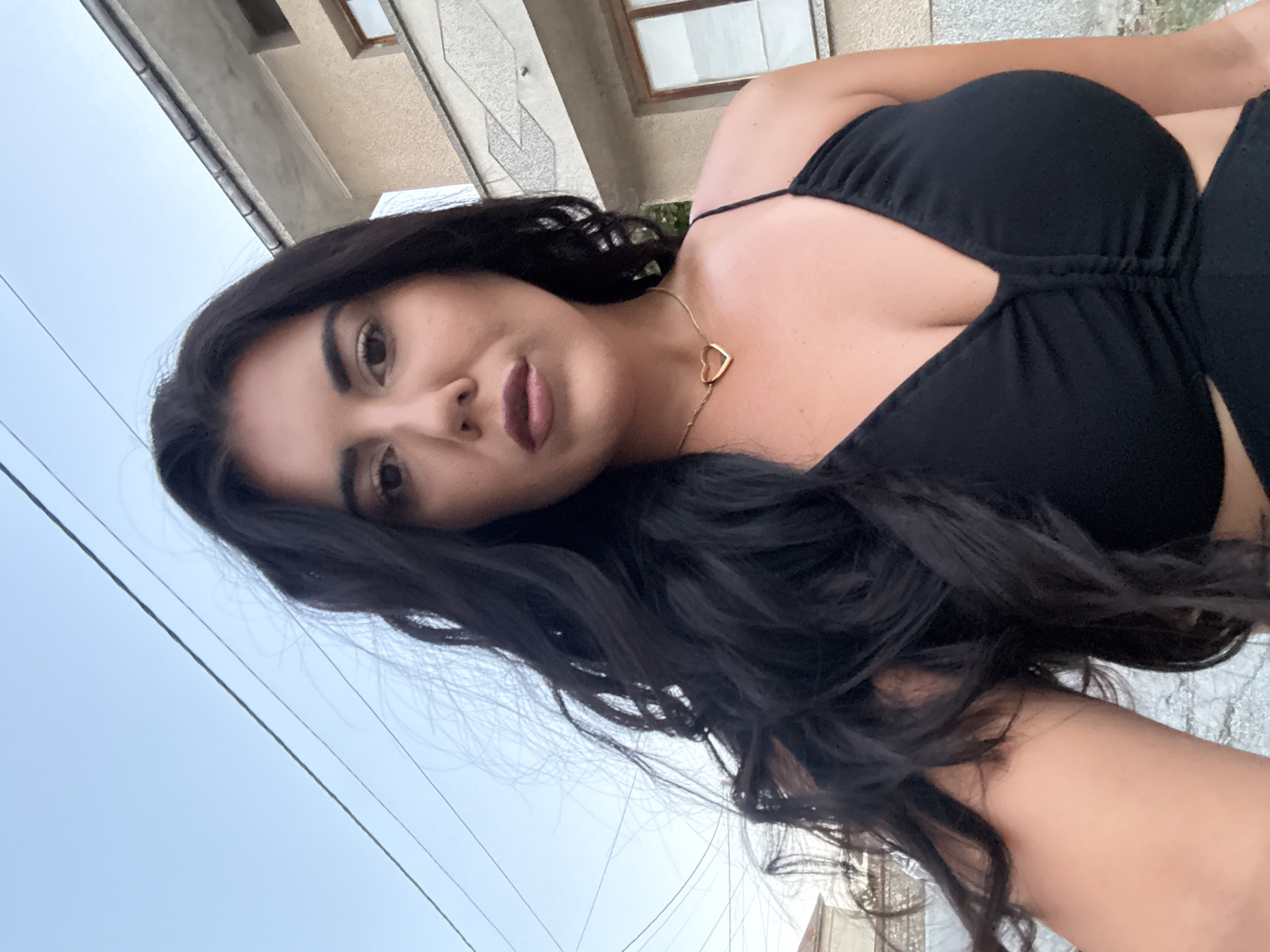 A selfie of a woman with long, dark, wavy hair is taken outdoors, with a building and power lines in the background. She is wearing a black dress with a heart-shaped necklace. The woman's face is partially visible, and she is looking directly at the camera. The sky is a clear blue. The building appears to be residential.